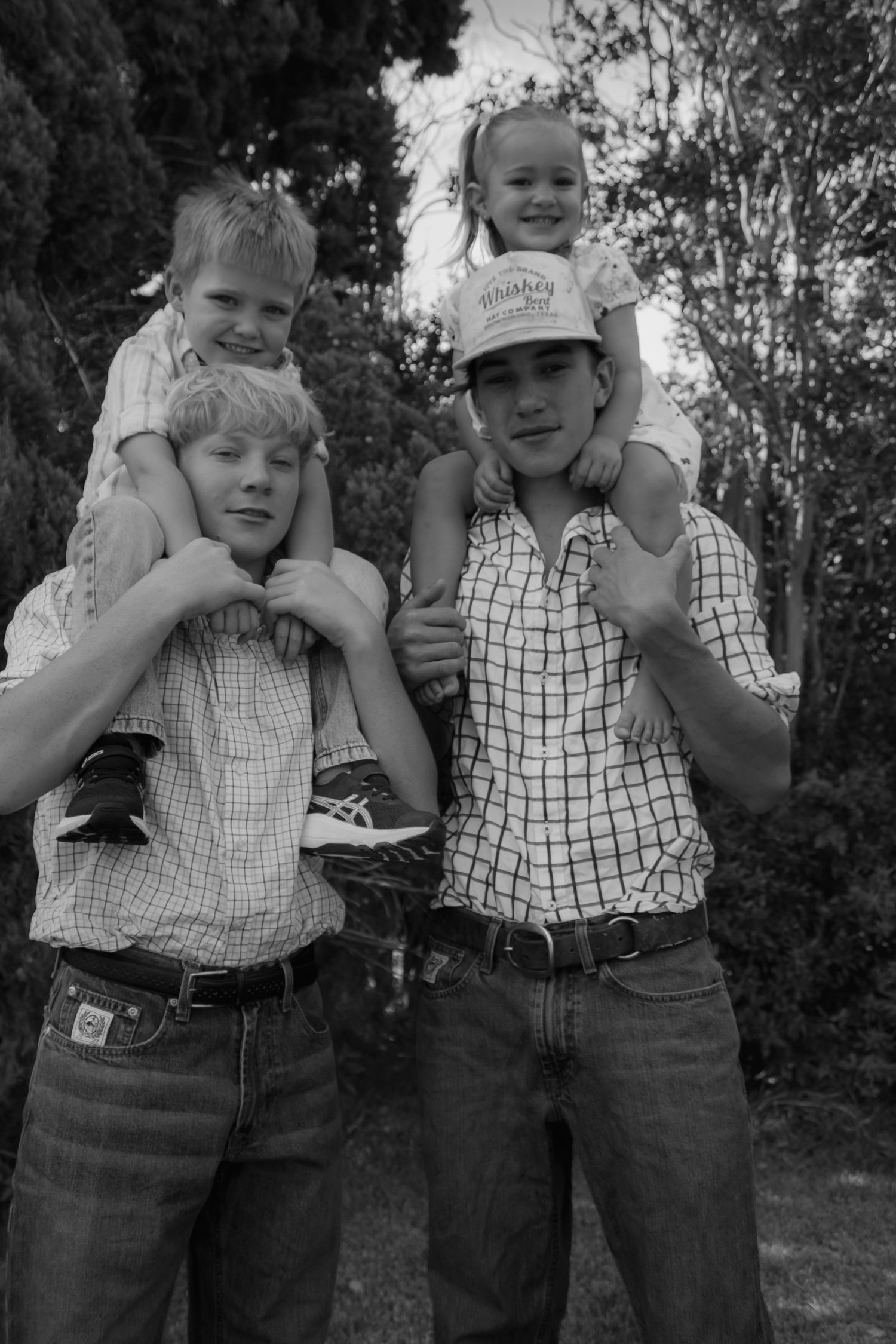 Black and white photo of two teenage boys each carrying a young child on their shoulders outdoors.