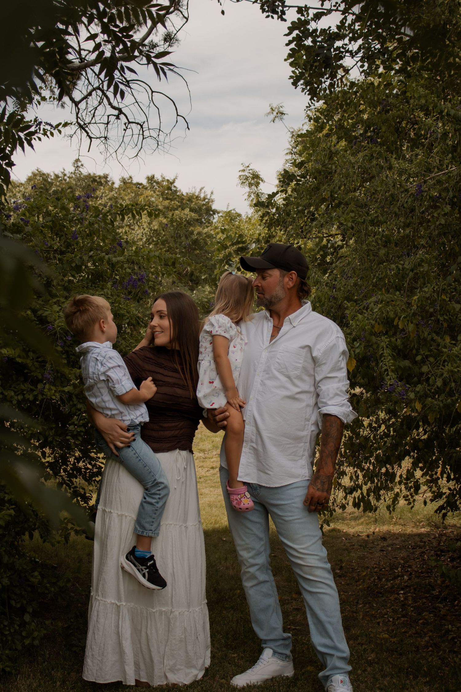 A family of four poses outdoors among lush green trees, parents holding two young children in a warm, candid moment.
