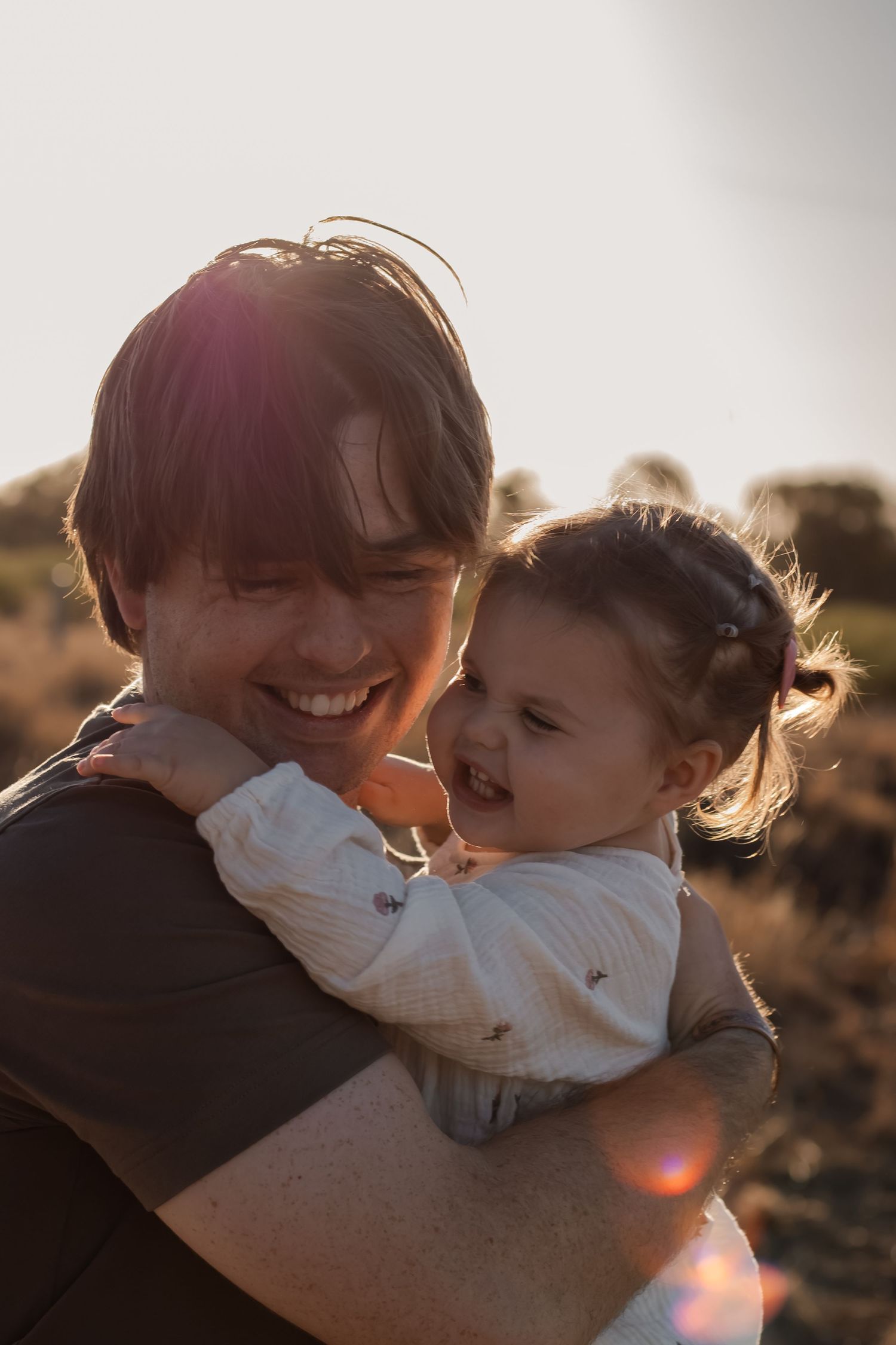 A smiling parent holds a laughing toddler girl in a warm golden sunset light outdoors.