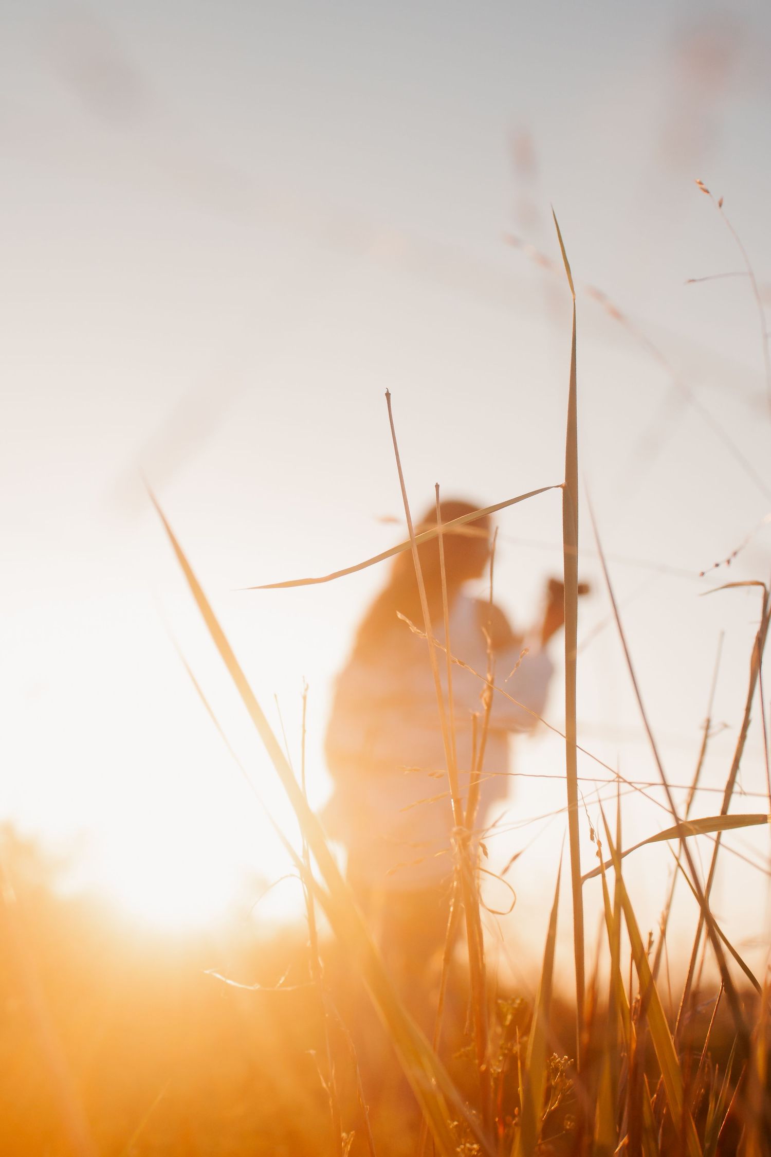 Silhouette of a woman standing in tall golden grass during a warm sunset, bathed in glowing orange light.