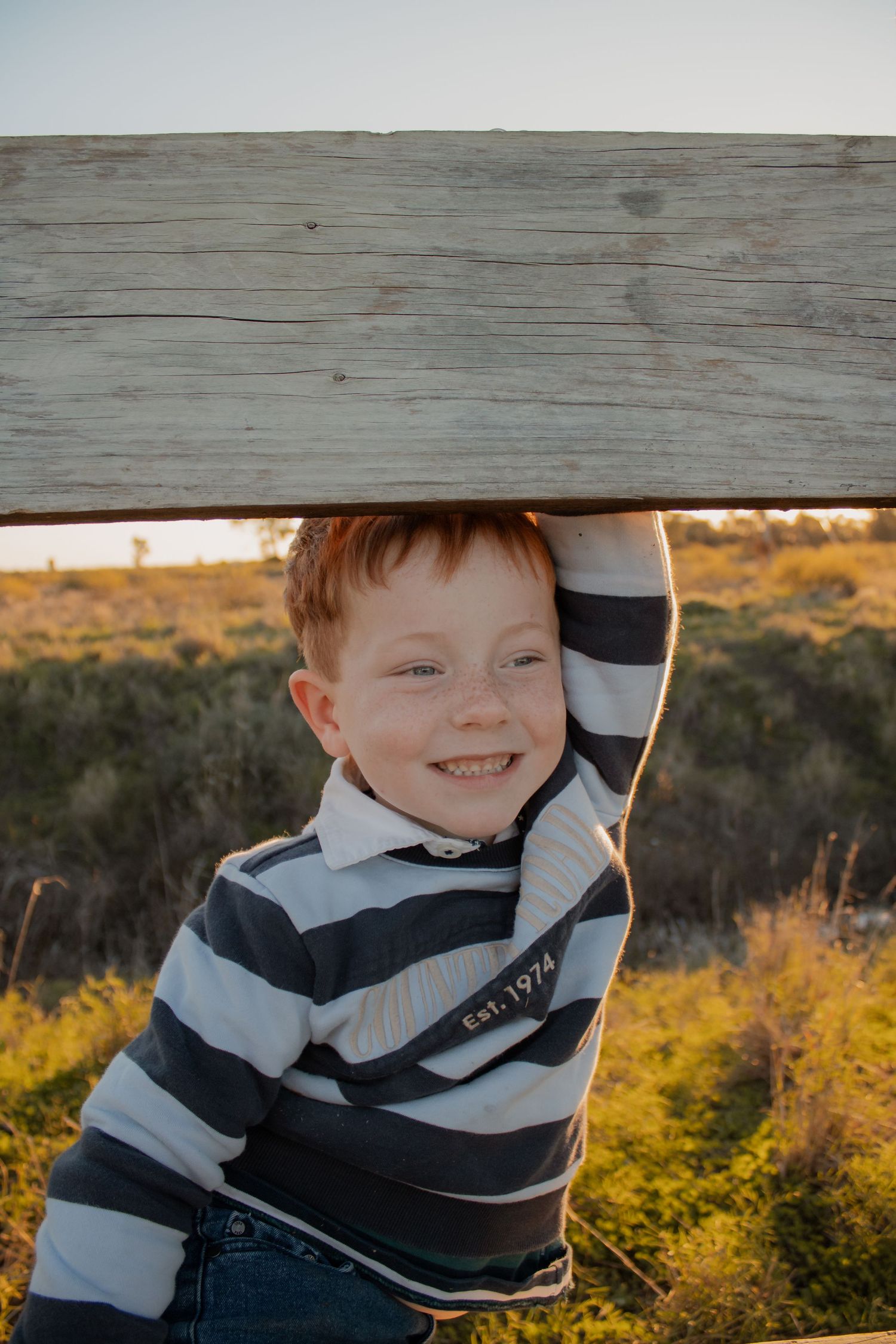 A smiling red-haired boy in a striped shirt leans against a wooden fence in a sunny outdoor field.