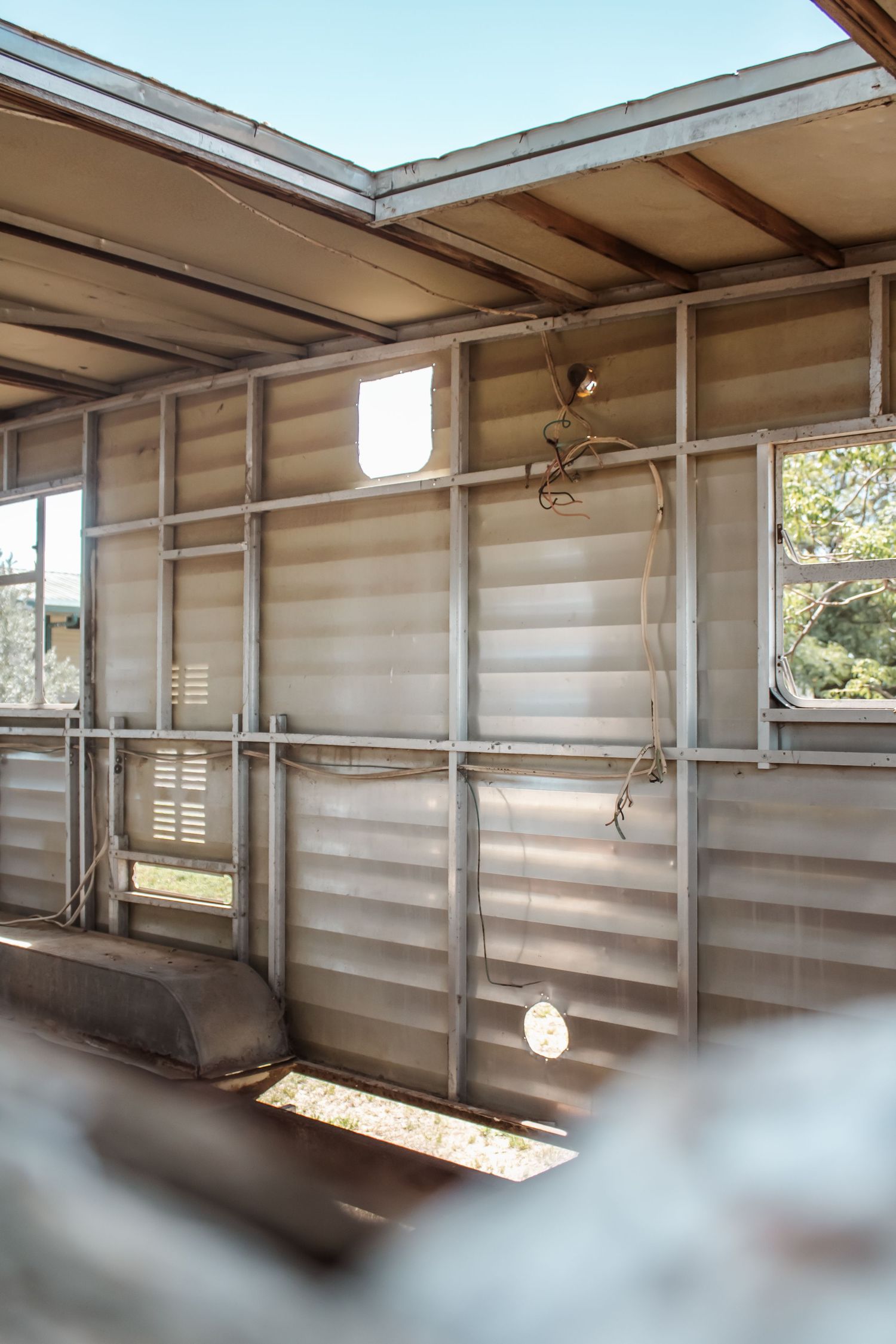 Sunlit abandoned caravan interior with corrugated metal walls, exposed framing, and dangling wires.