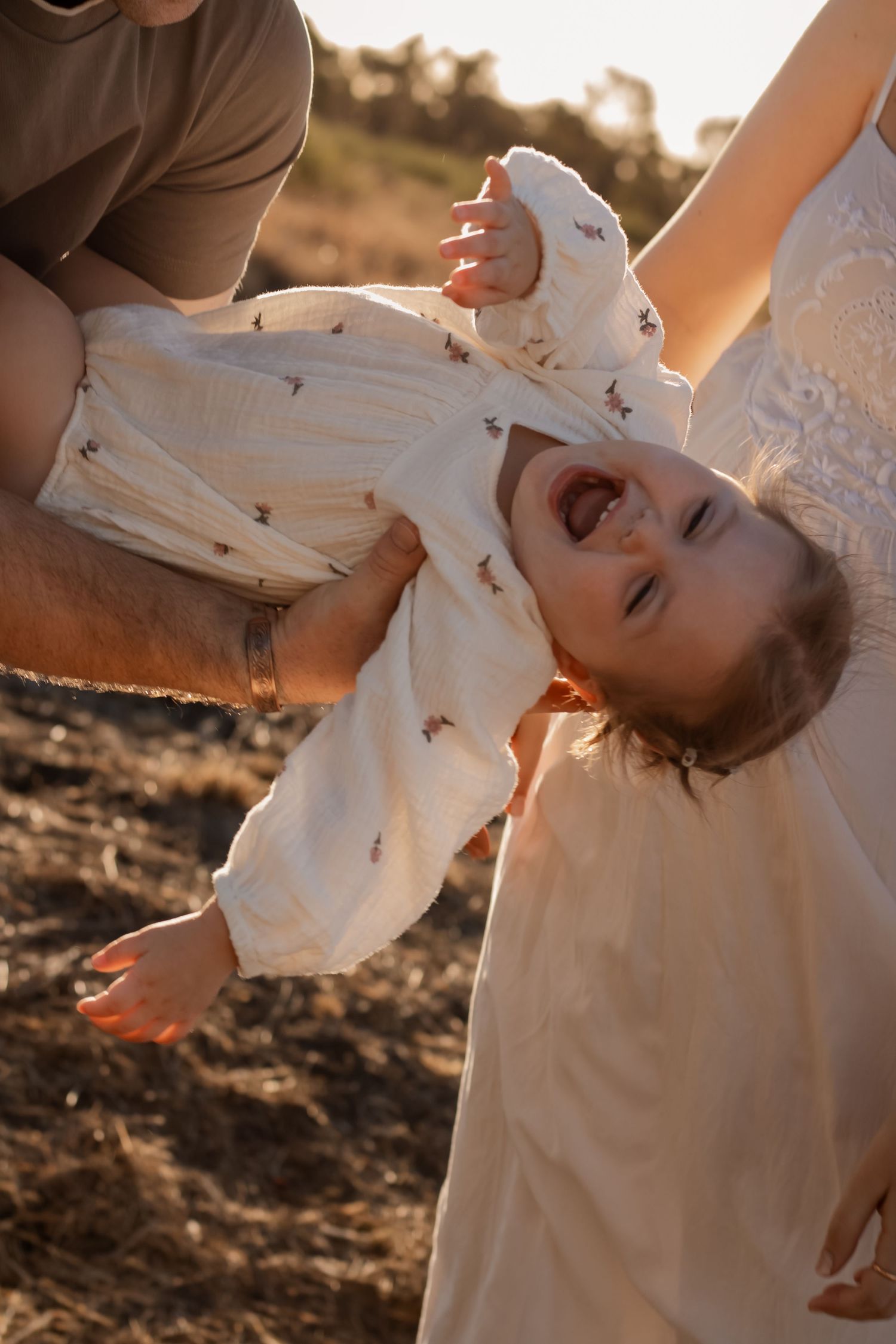 Parents playfully flip laughing toddler upside down during golden hour outdoor family photo session.