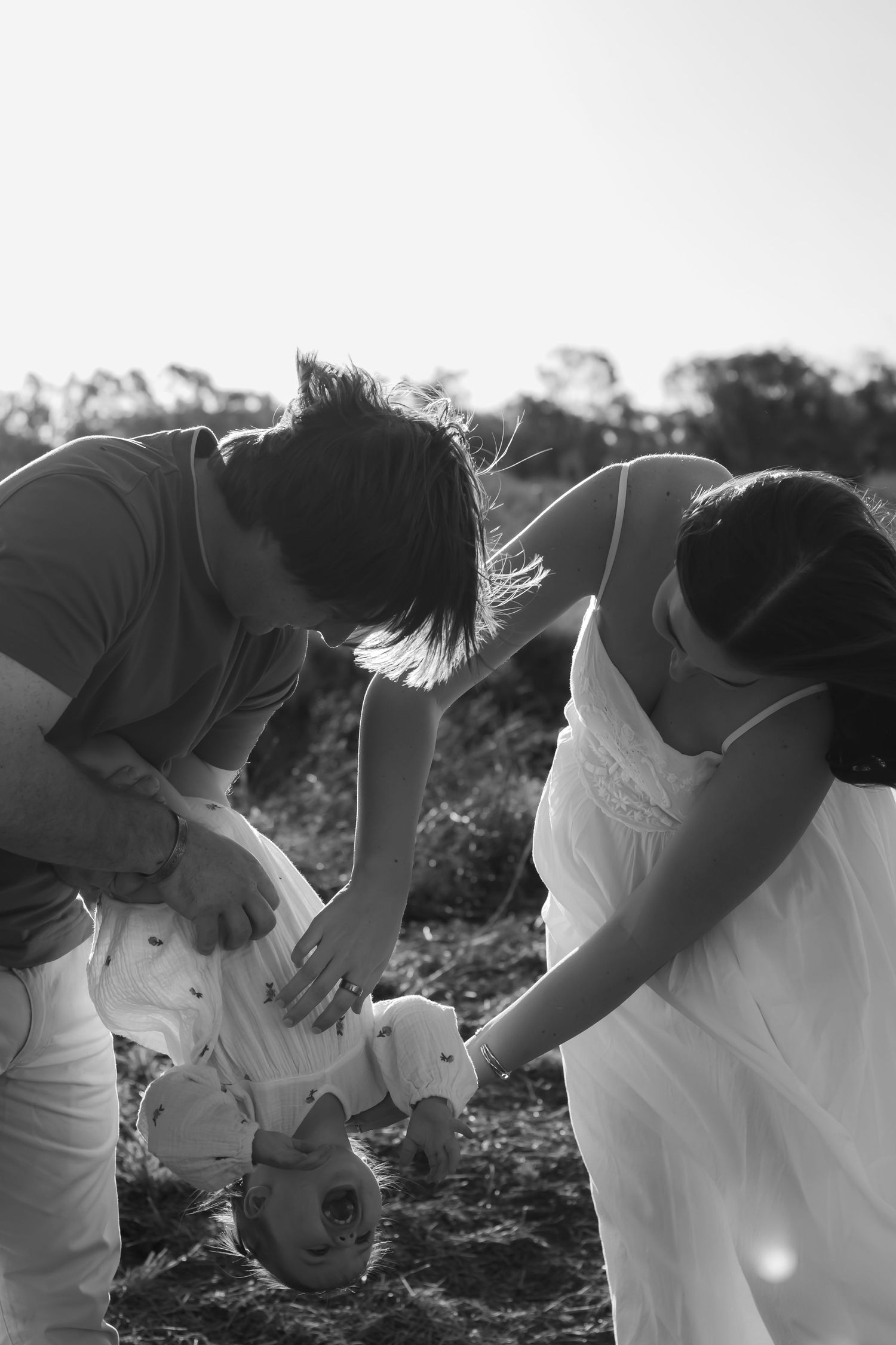 Black and white photo of two parents bending down to tend to their baby outdoors in a natural setting.