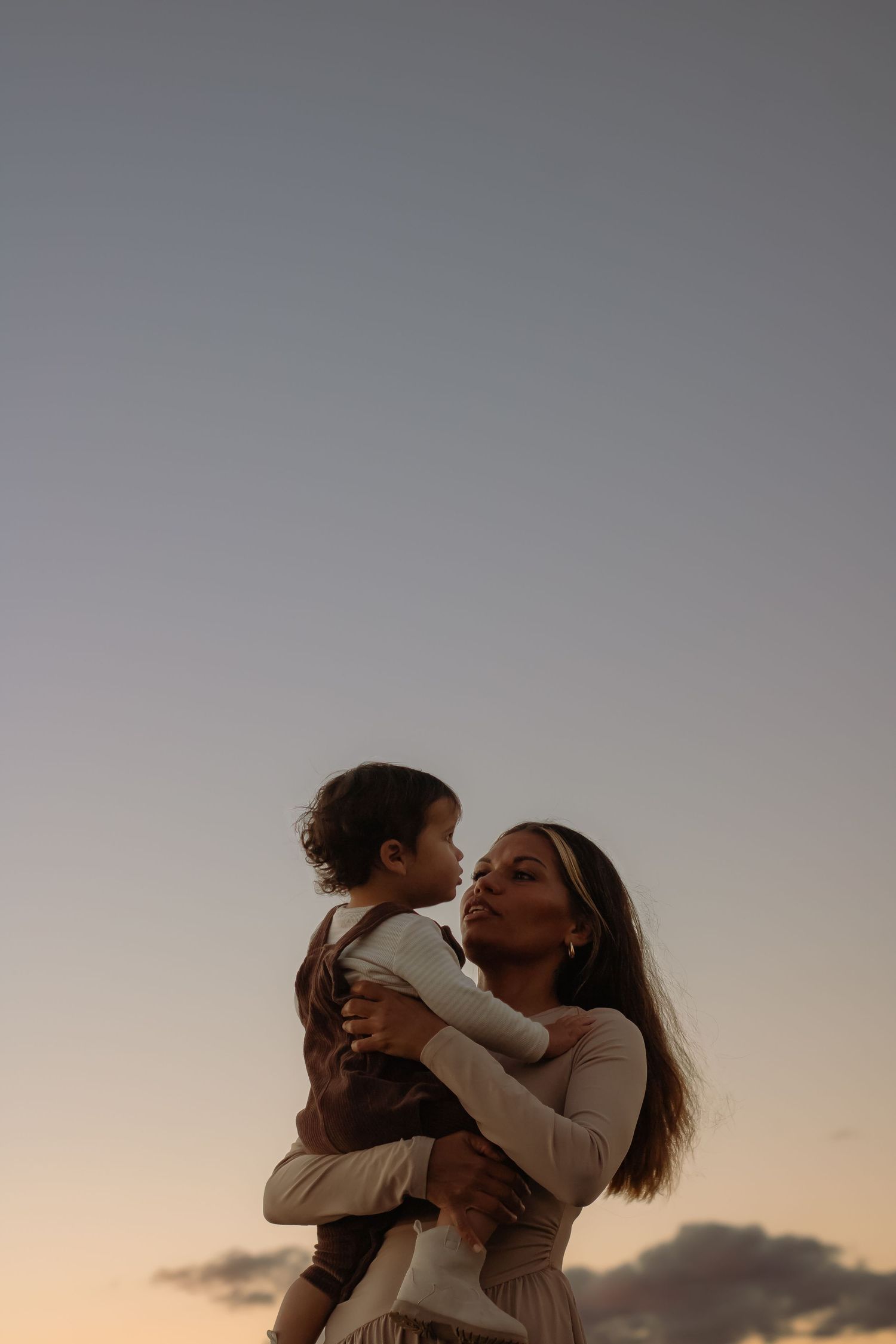 Mother holding young toddler against a soft pastel sunset sky, sharing a loving gaze.