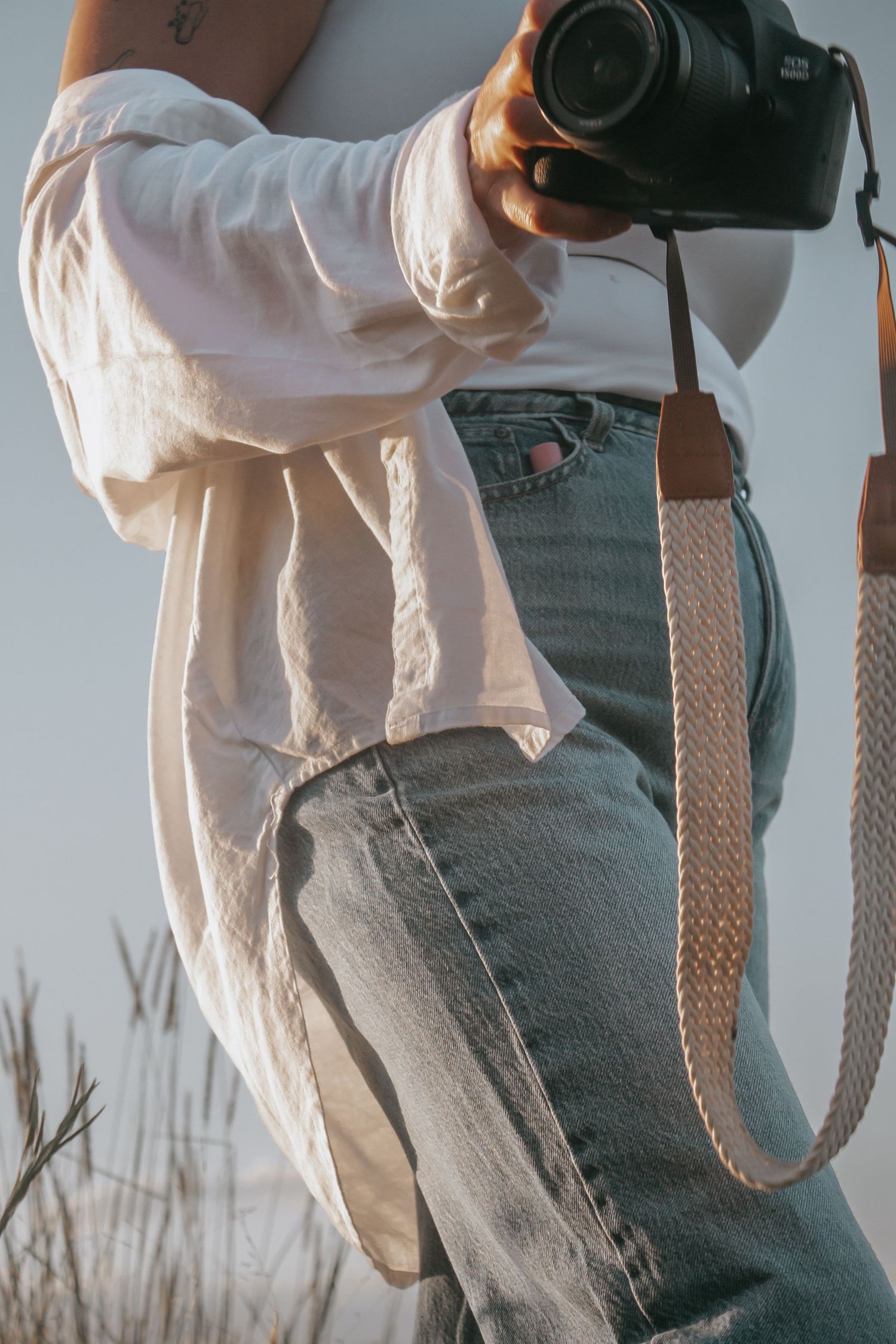 Close-up of woman in white shirt and jeans holding DSLR camera with braided strap.