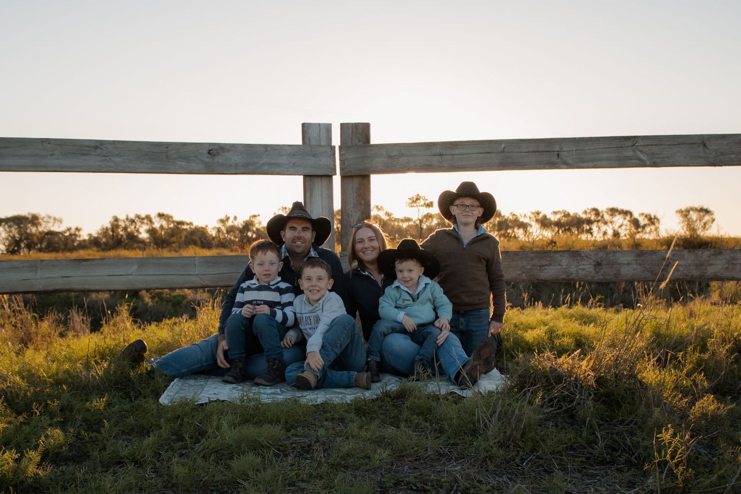 Family of six posing outdoors at golden hour near a wooden fence in a rural countryside setting.