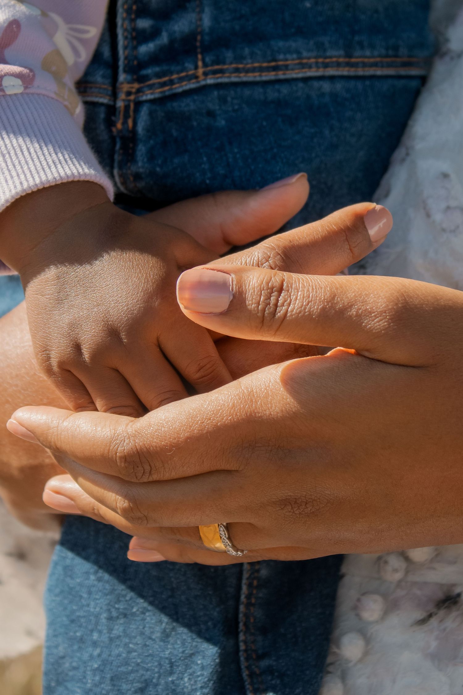 Close-up of adult hands gently holding a small child's hand, with a gold ring visible outdoors in warm sunlight.