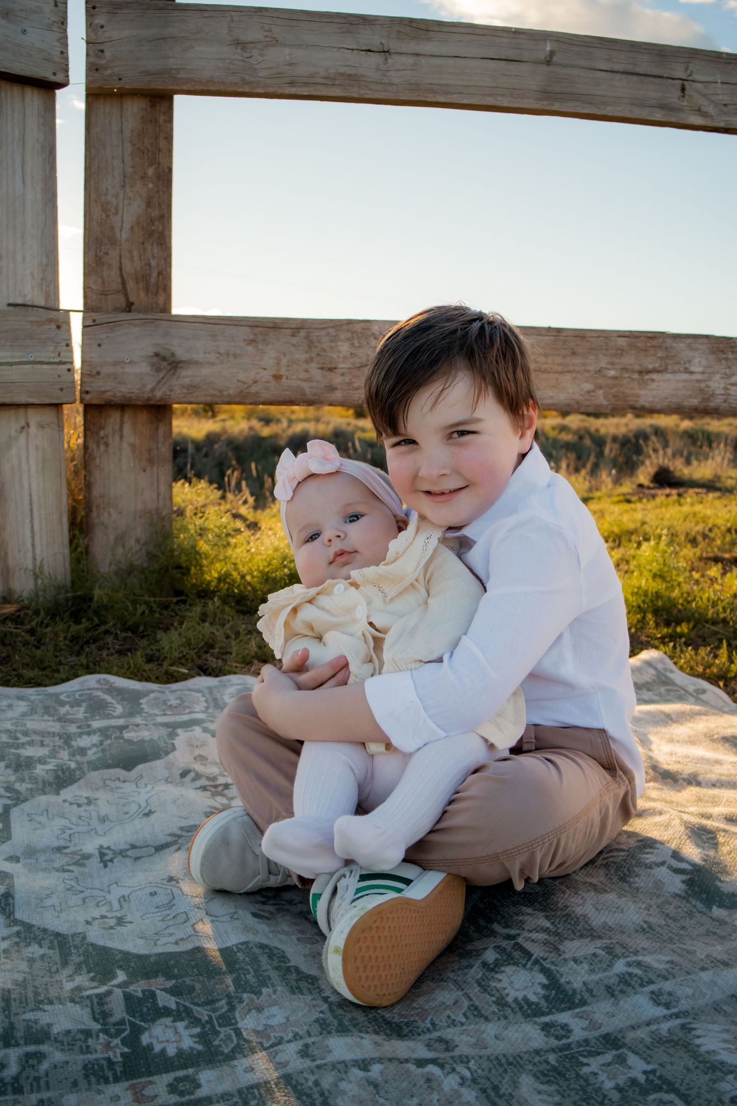 Young boy smiling and holding baby sister on a blanket outdoors near a wooden fence at golden hour.