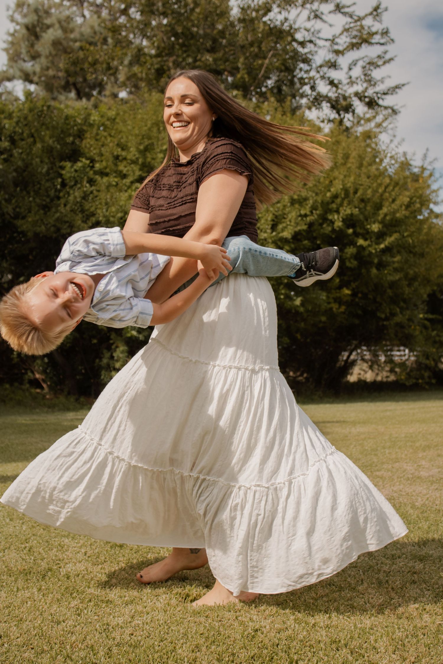 A woman in a white tiered skirt joyfully lifts a child outdoors on green grass with trees behind.