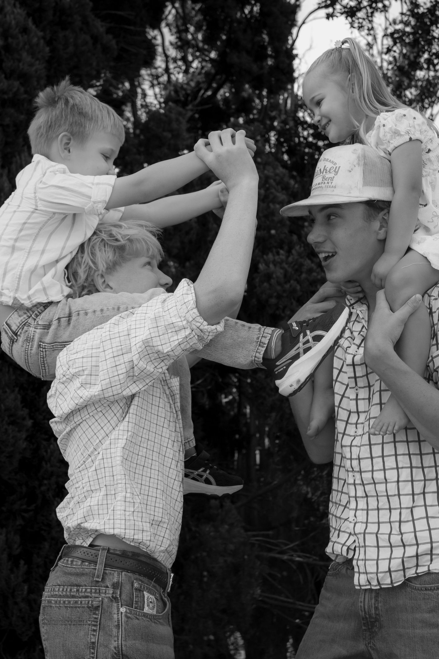 Black and white photo of two teens carrying young children on their shoulders, playing outdoors near trees.