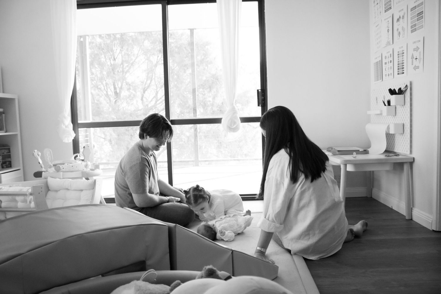 Black and white photo of a family with a dog relaxing together in a bright, cozy bedroom near a large window.
