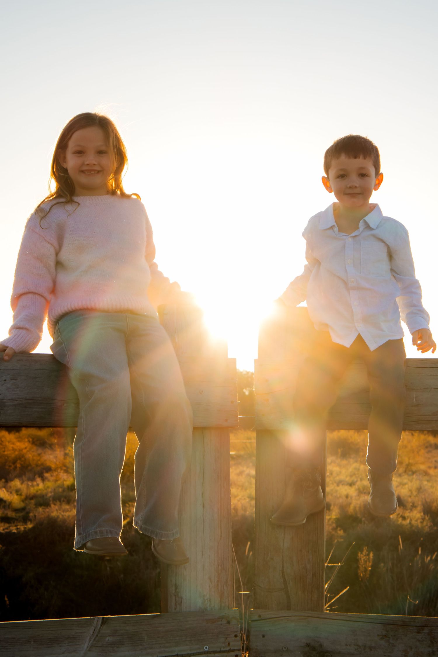Two children sitting on a wooden fence at golden sunset, girl in pink sweater and boy in white shirt smiling outdoors.