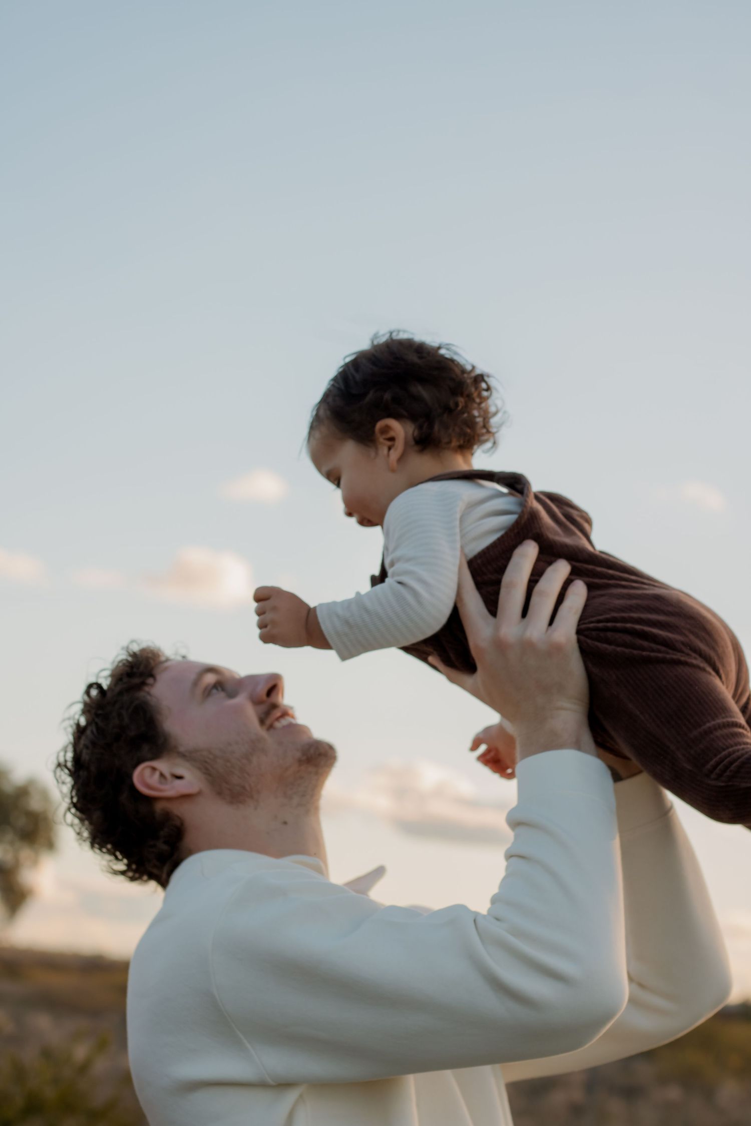 A smiling man in a white sweater lifts a curly-haired toddler in brown overalls against a soft sky.