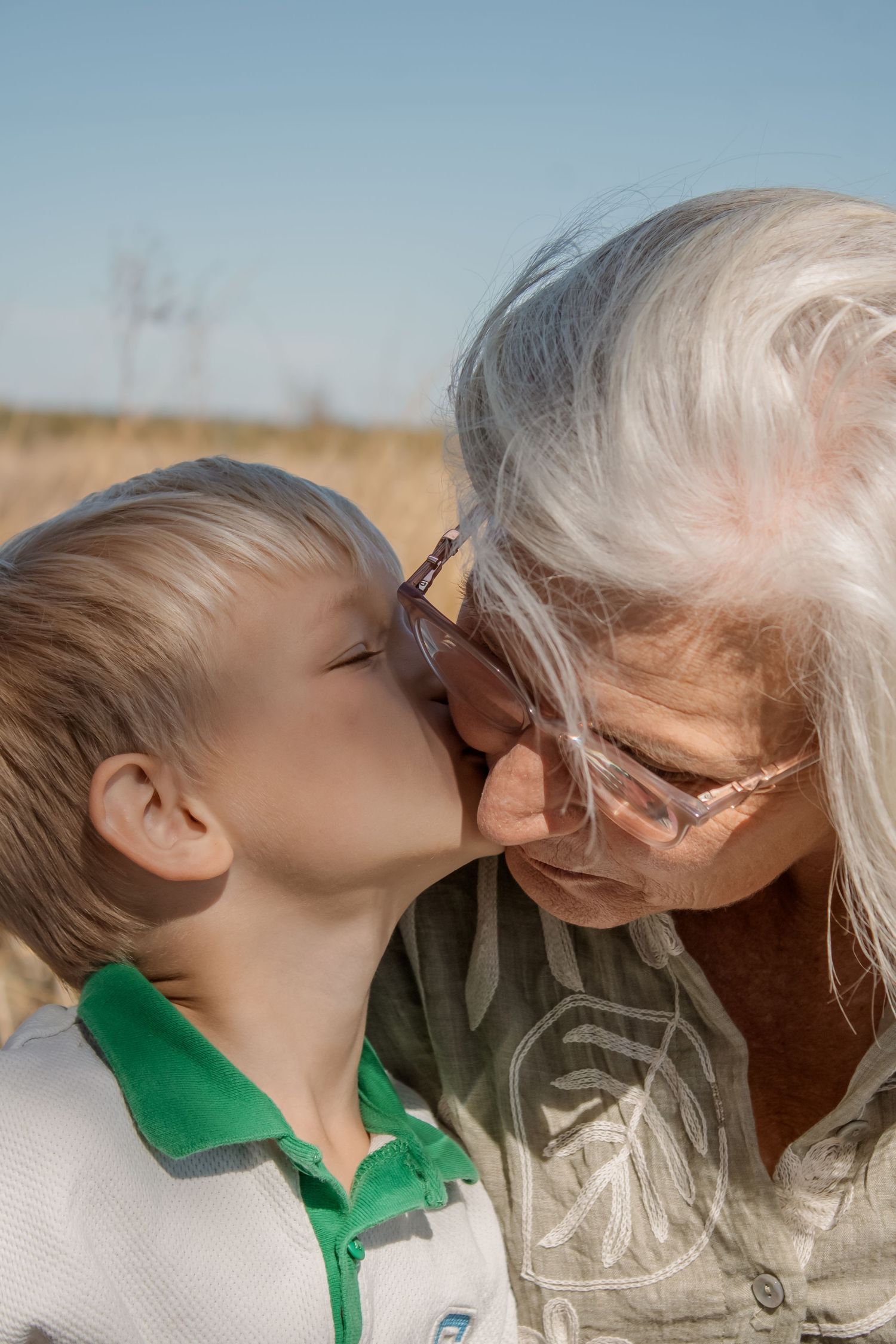 Young boy kissing elderly grandmother on the cheek outdoors in a sunny field, showing family love.