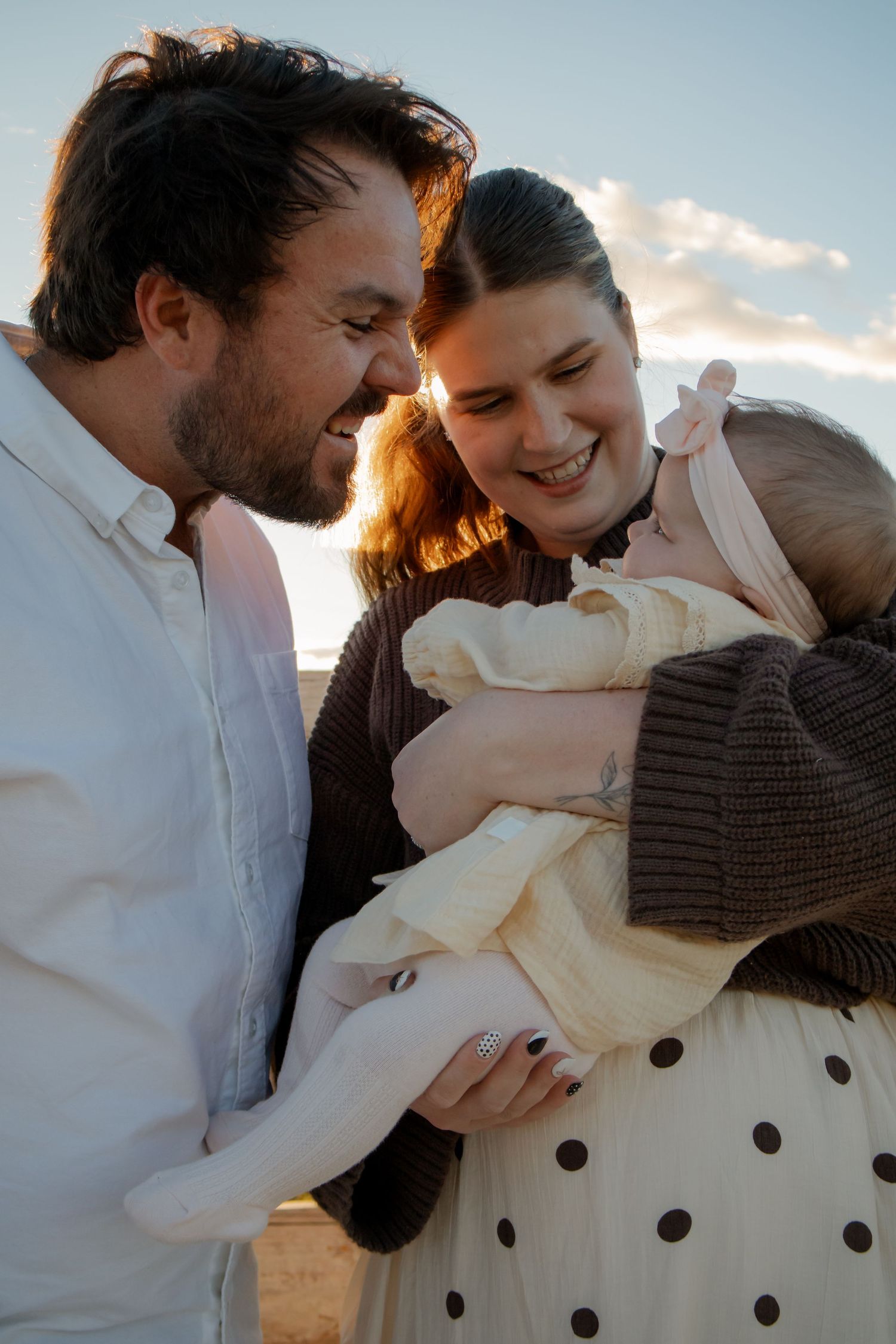 A couple smiles lovingly at their baby outdoors during golden hour sunset light.