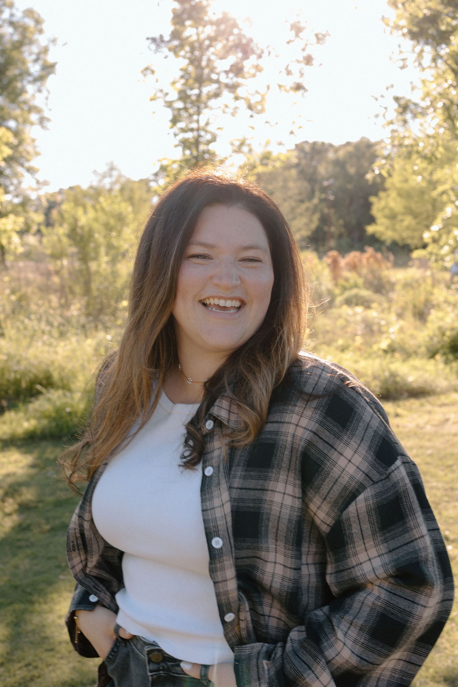 A young woman in a plaid shirt laughs joyfully outdoors in a bright sunlit green setting.