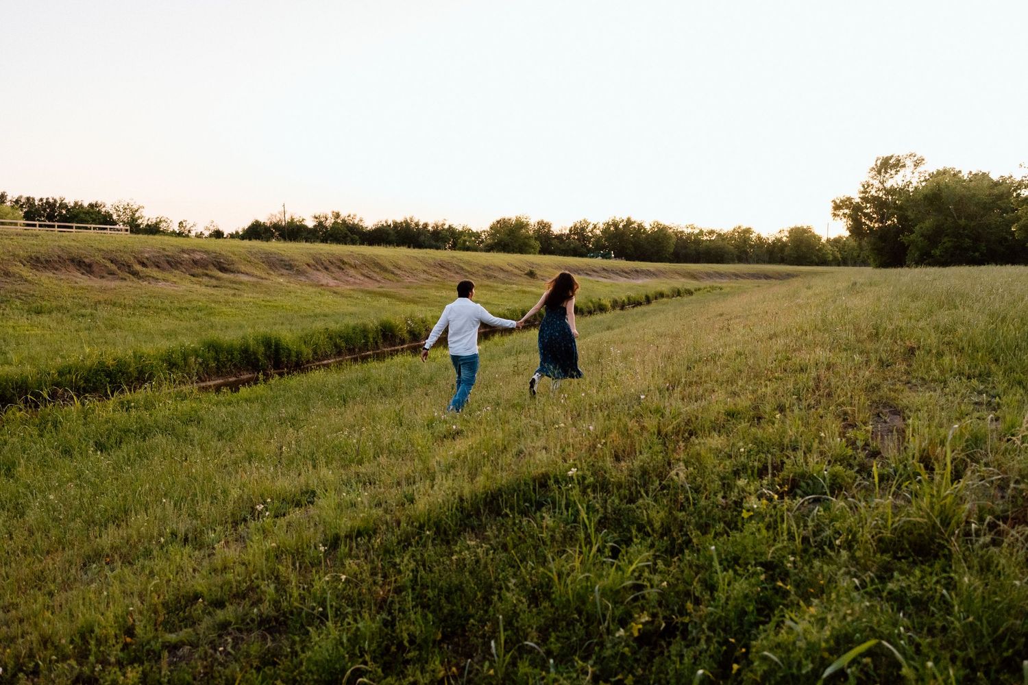 Couple holding hands while walking through a scenic green meadow at golden hour sunset.