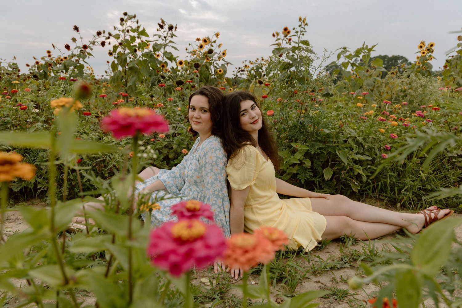 Two young women in summer dresses sit back-to-back in a vibrant wildflower garden.