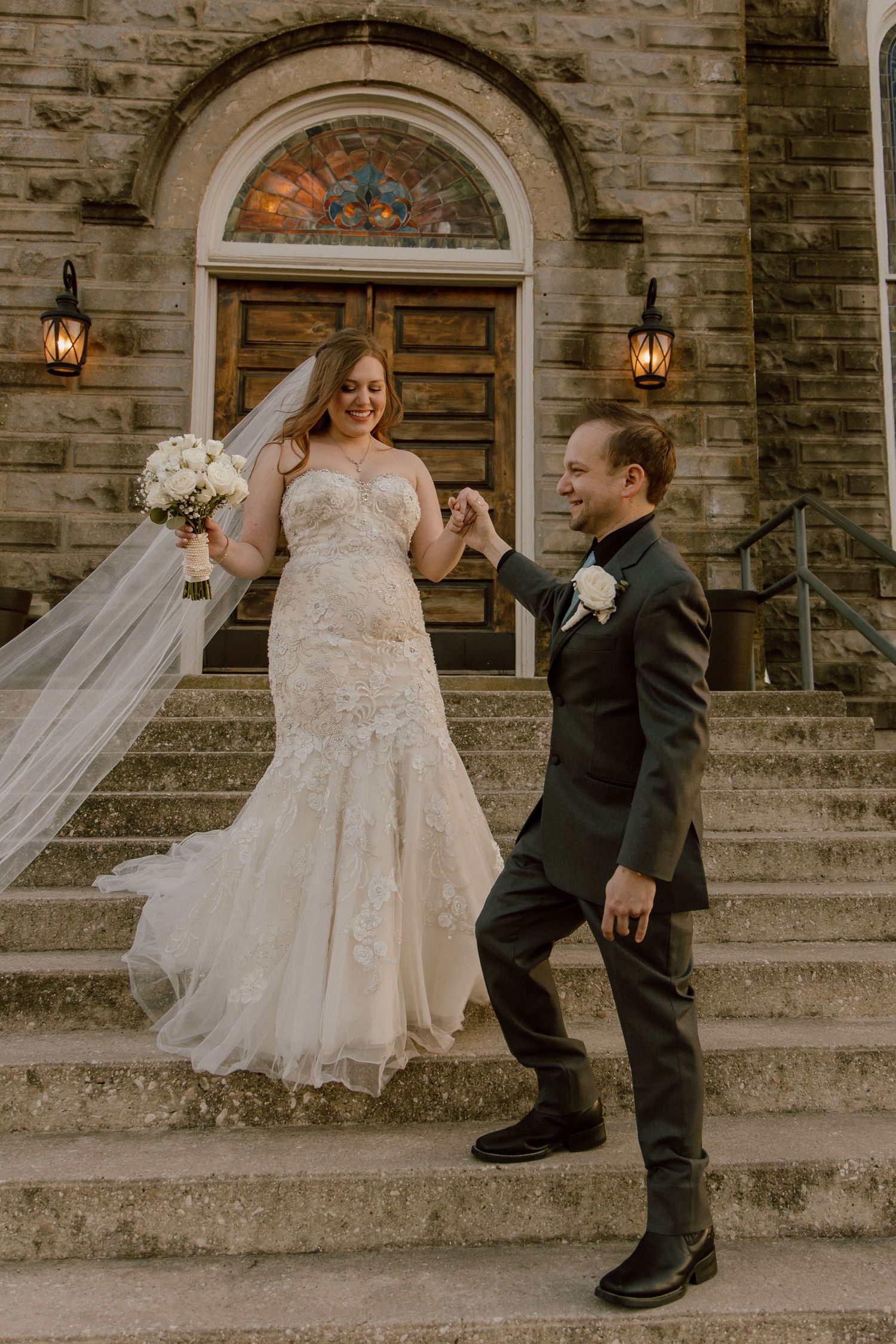 Bride and groom pose on stone church steps, bride holding a white bouquet.