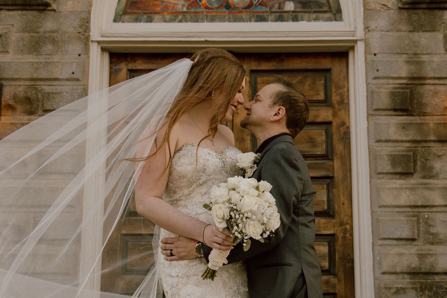 Bride and groom sharing a tender kiss with flowing veil in front of a rustic wooden door.