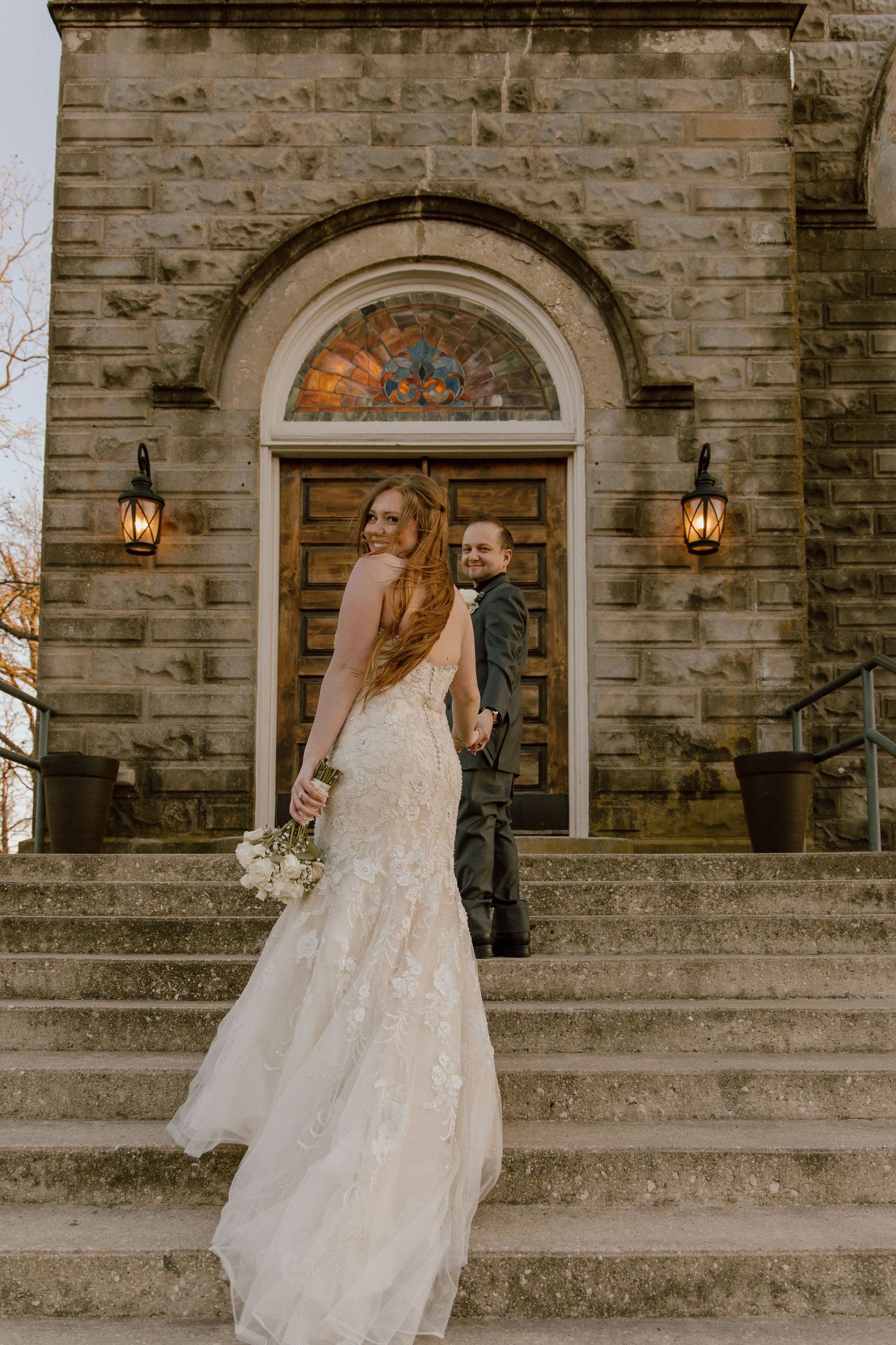Bride in lace gown and groom stand on stone church steps holding hands outdoors.
