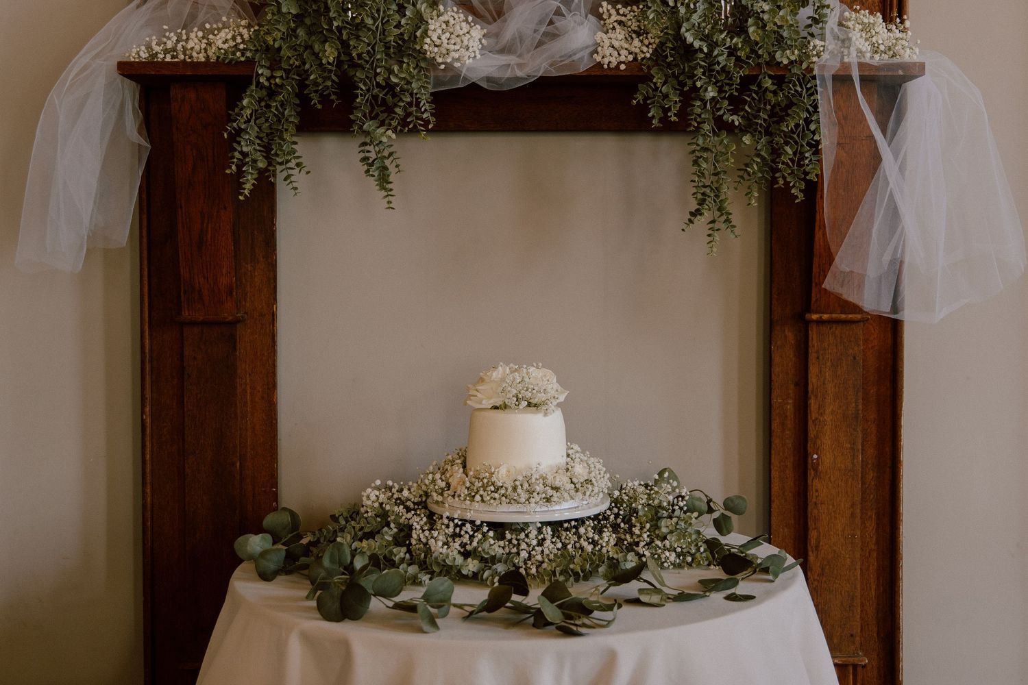 Single-tier white wedding cake decorated with baby's breath under wooden arch with greenery.