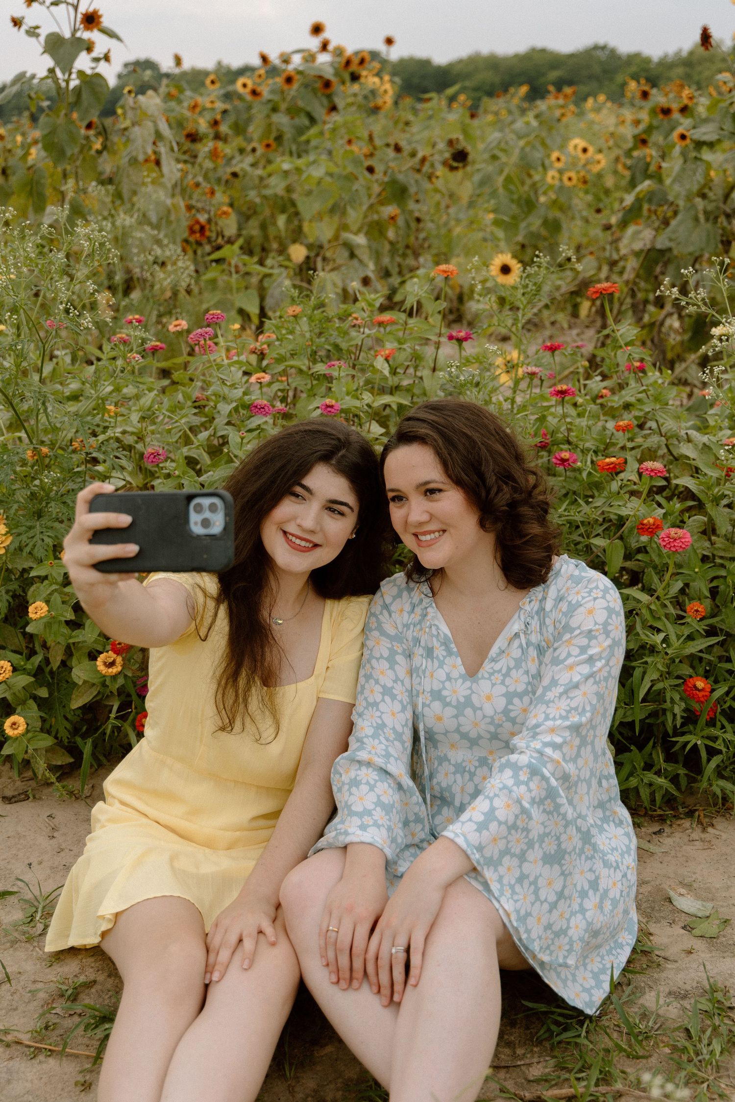 Two young women in floral dresses take a selfie in a colorful sunflower and zinnia field.