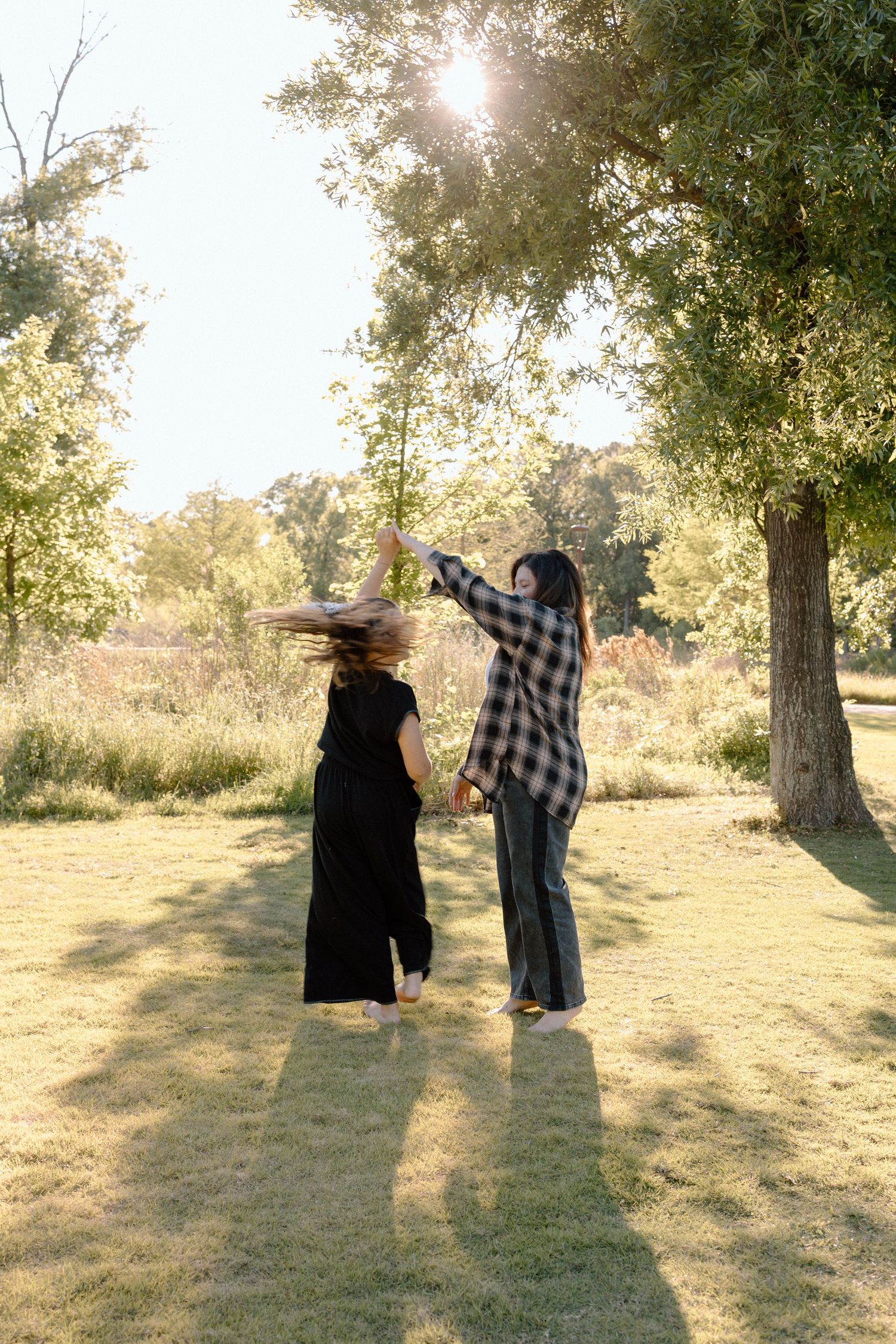 A mother and daughter dance and spin together in a sunny green park near a large tree.