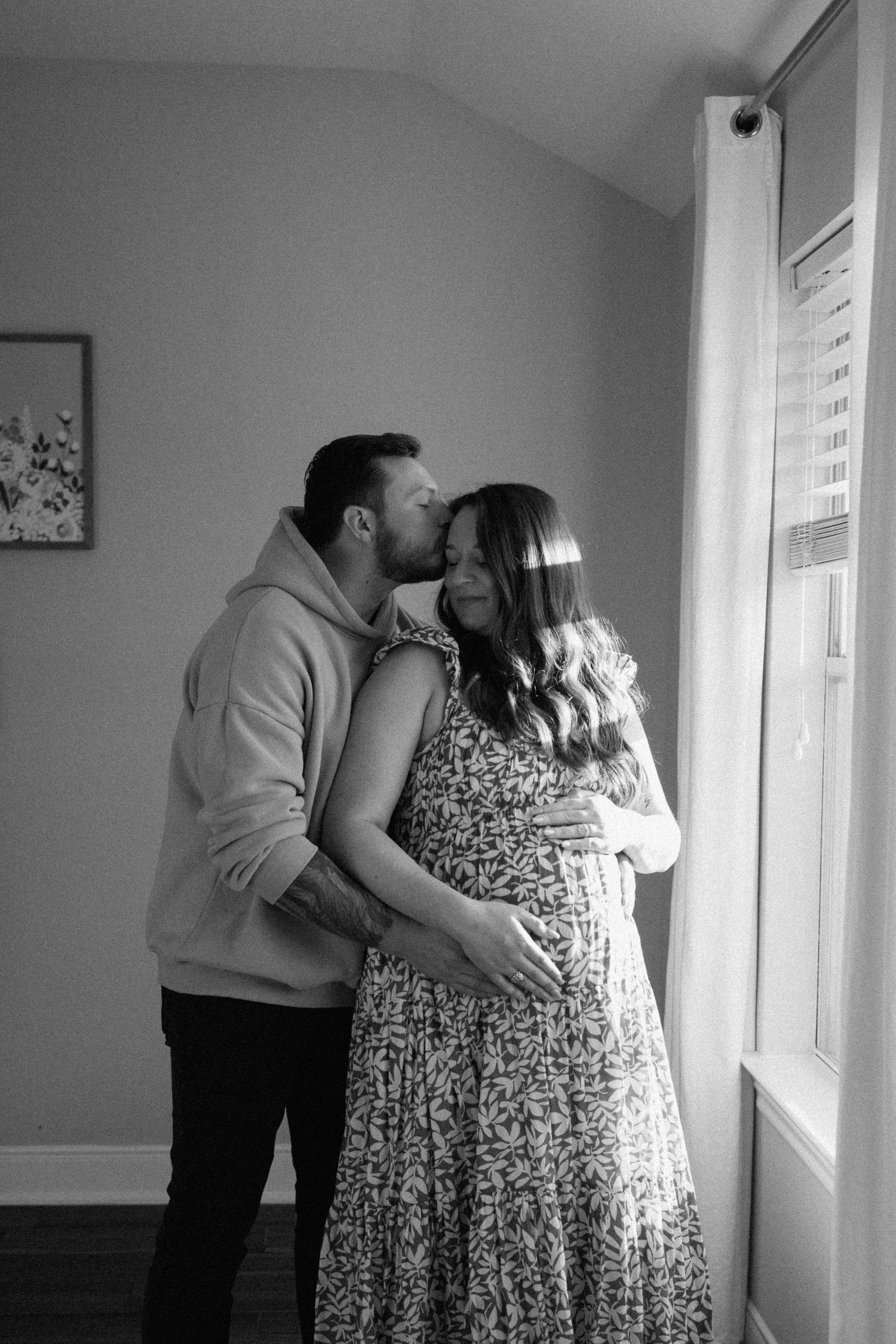 Black and white maternity photo of couple embracing near a window, man kissing womans head.