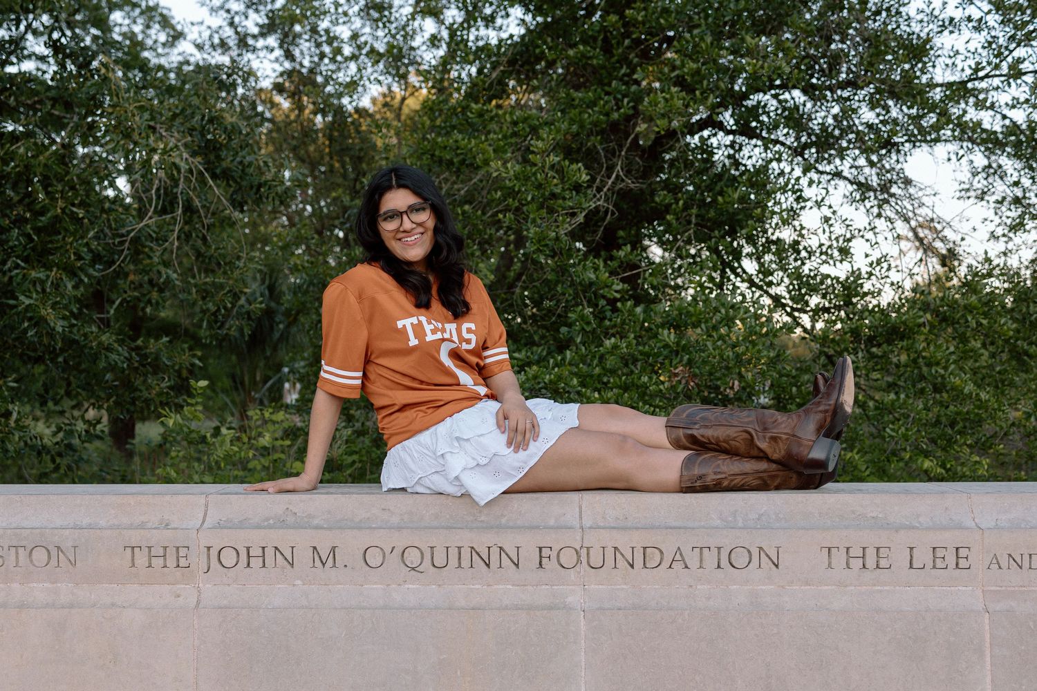 Young woman in Texas jersey and cowboy boots sitting on stone wall with John M. O'Quinn Foundation engraving.