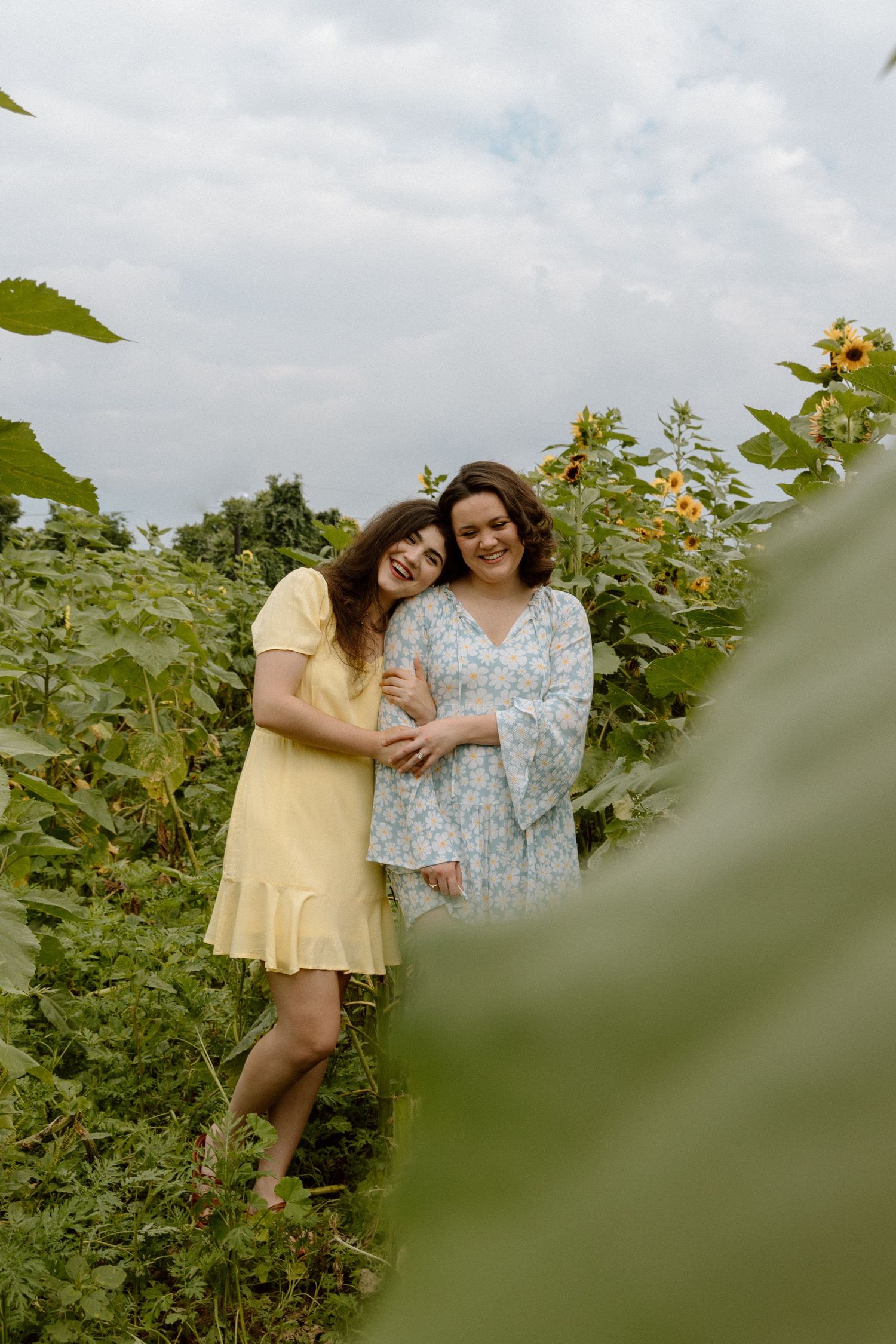 Two women in summer dresses laughing and embracing among tall sunflowers under an overcast sky.