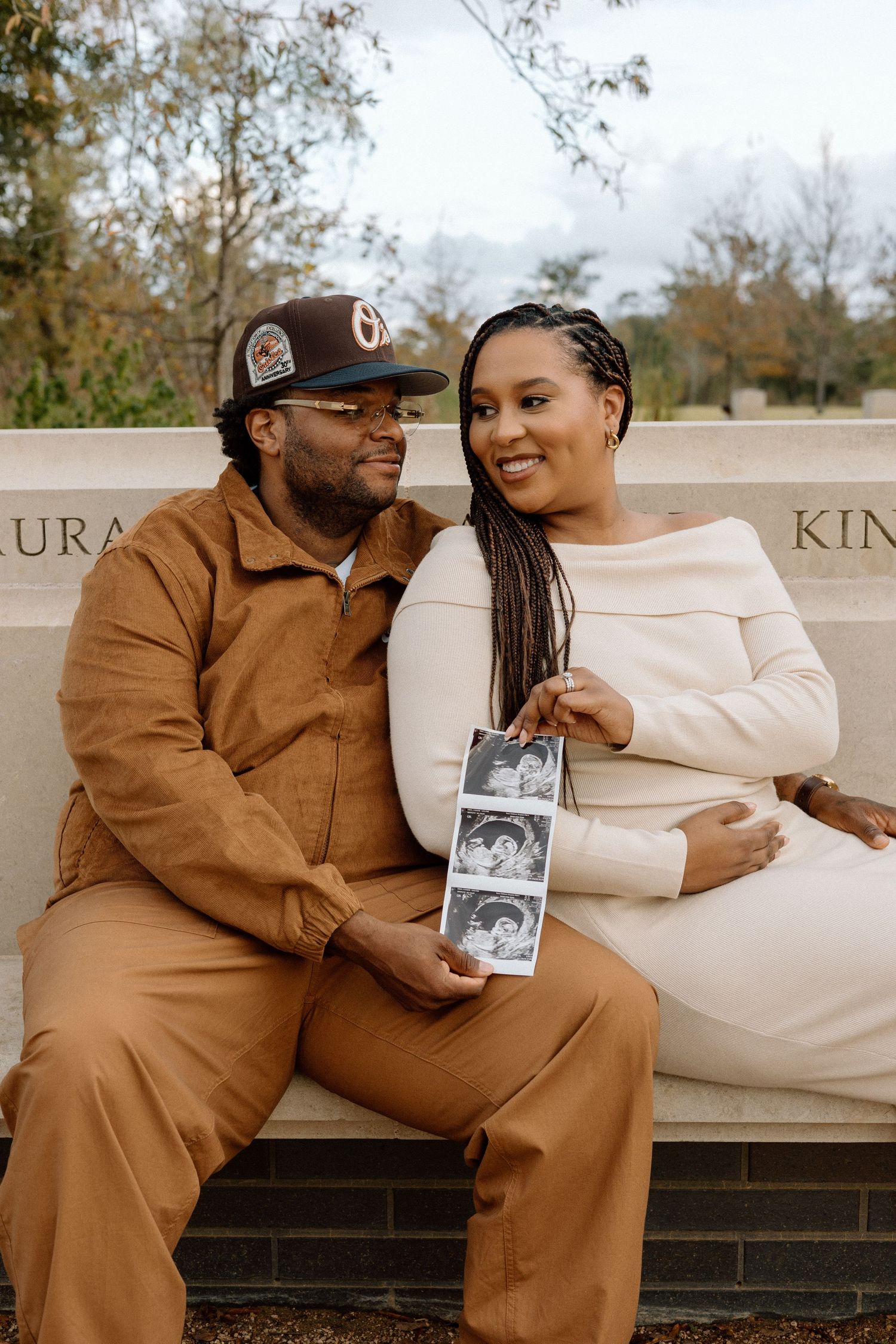 Couple sitting on bench holding ultrasound photos during outdoor maternity portrait session in park.