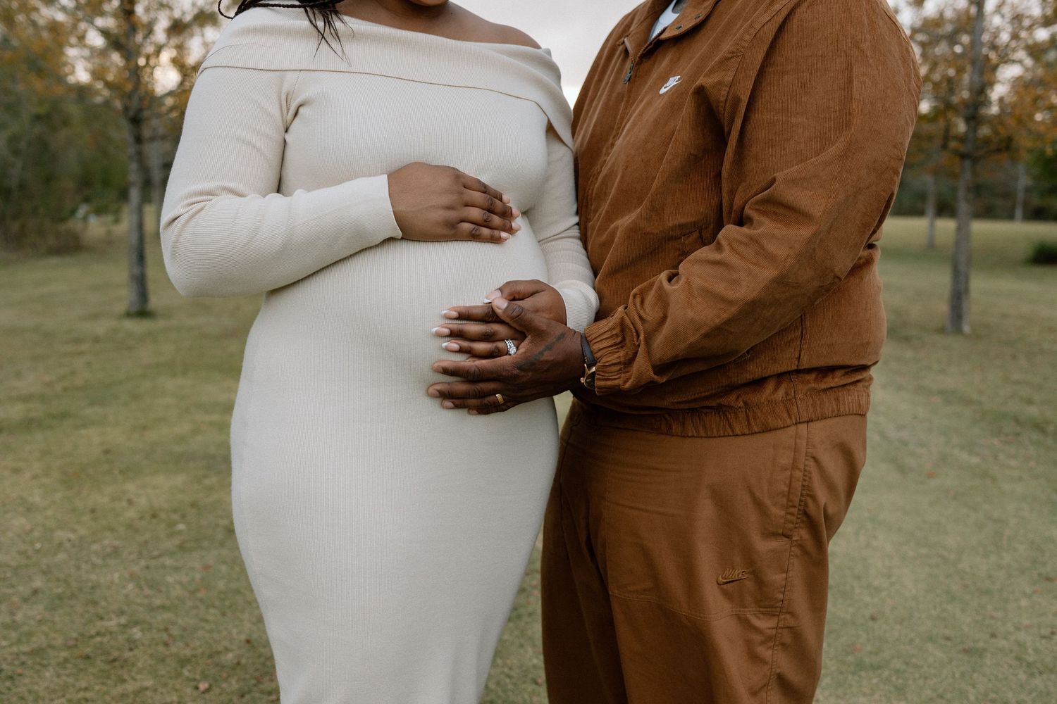 Expecting couple in neutral tones gently cradling baby bump during outdoor maternity photoshoot.