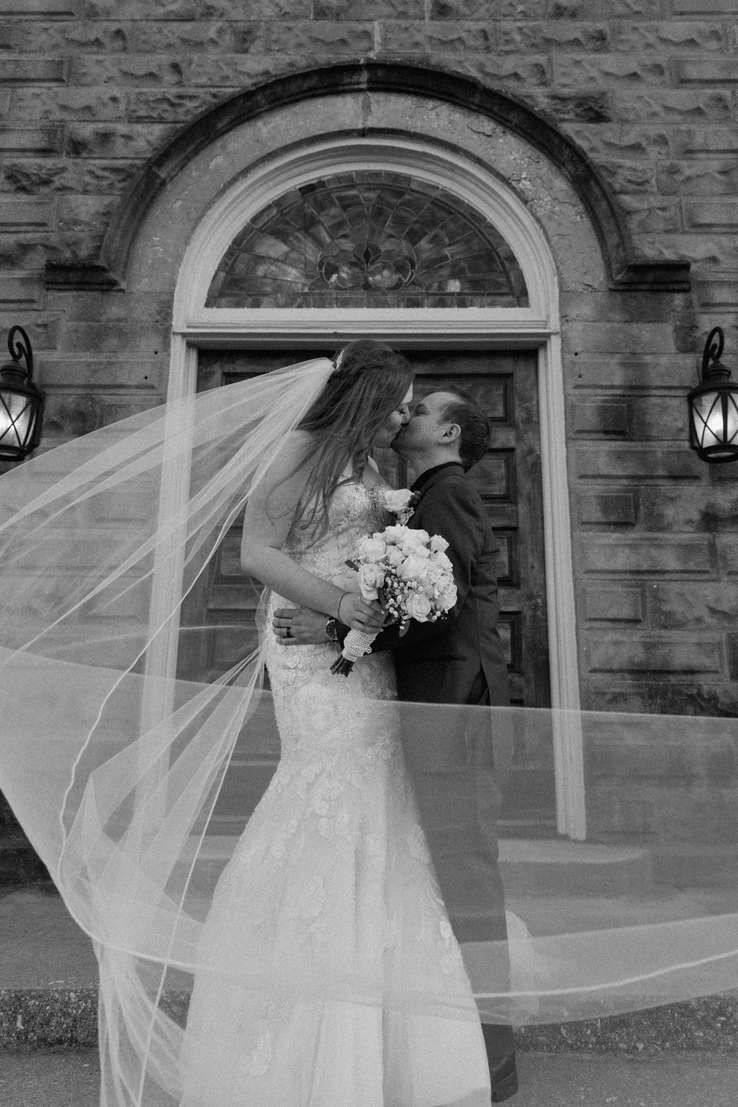 Black and white wedding photo of couple kissing outside a stone church with veil flowing.