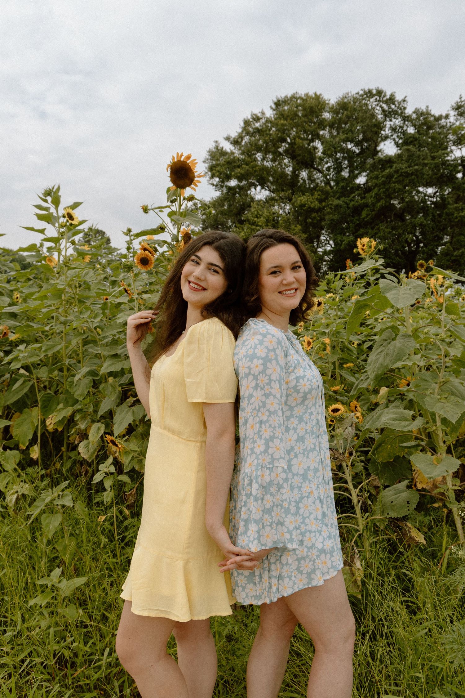 Two young women in floral summer dresses smiling back to back in a sunflower field.