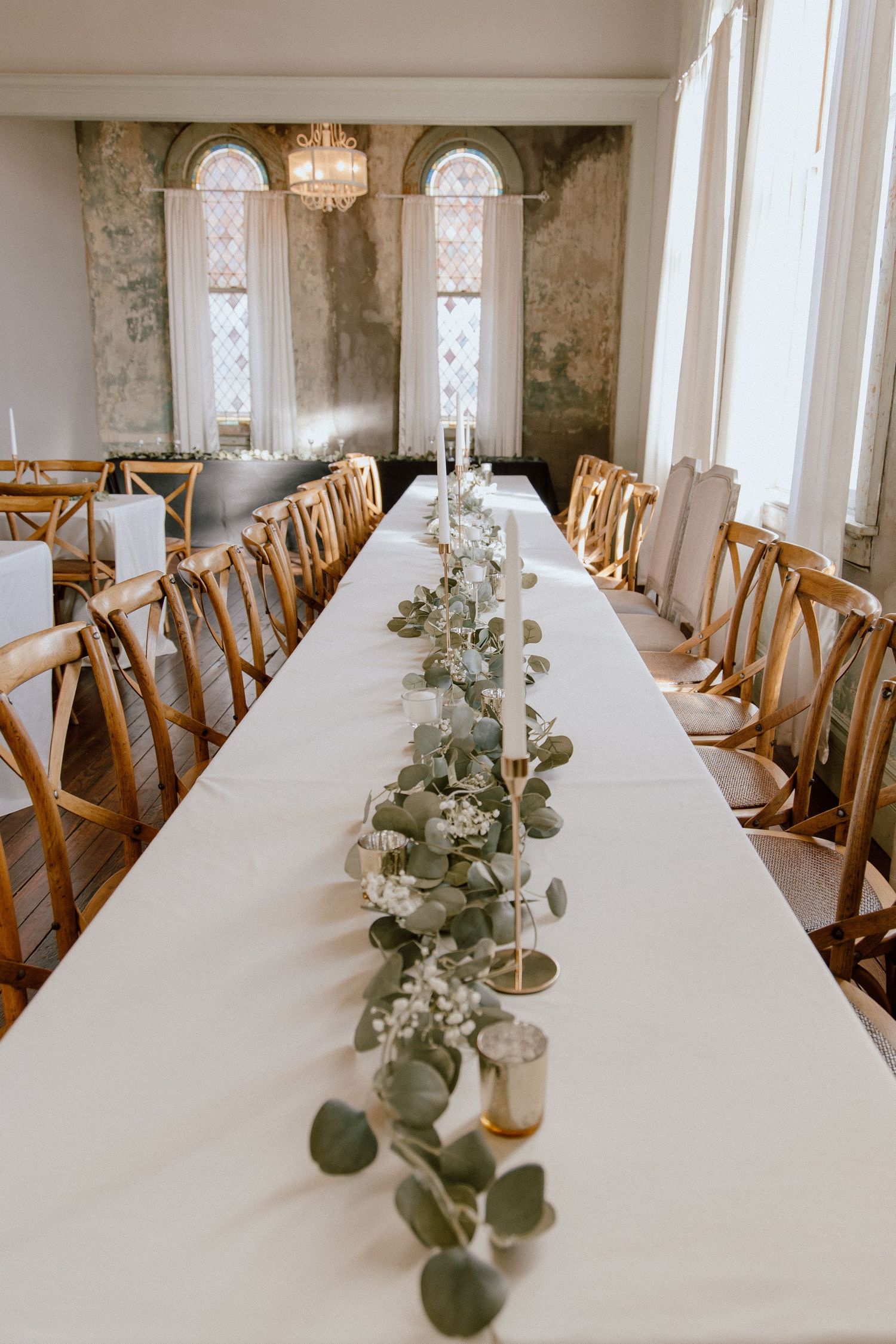 Elegant wedding reception table with eucalyptus garland runner, gold candle holders, and cross-back chairs.
