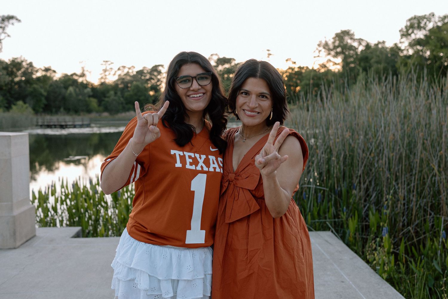 Two women wearing University of Texas orange jerseys making Longhorns hand signs outdoors near a pond.