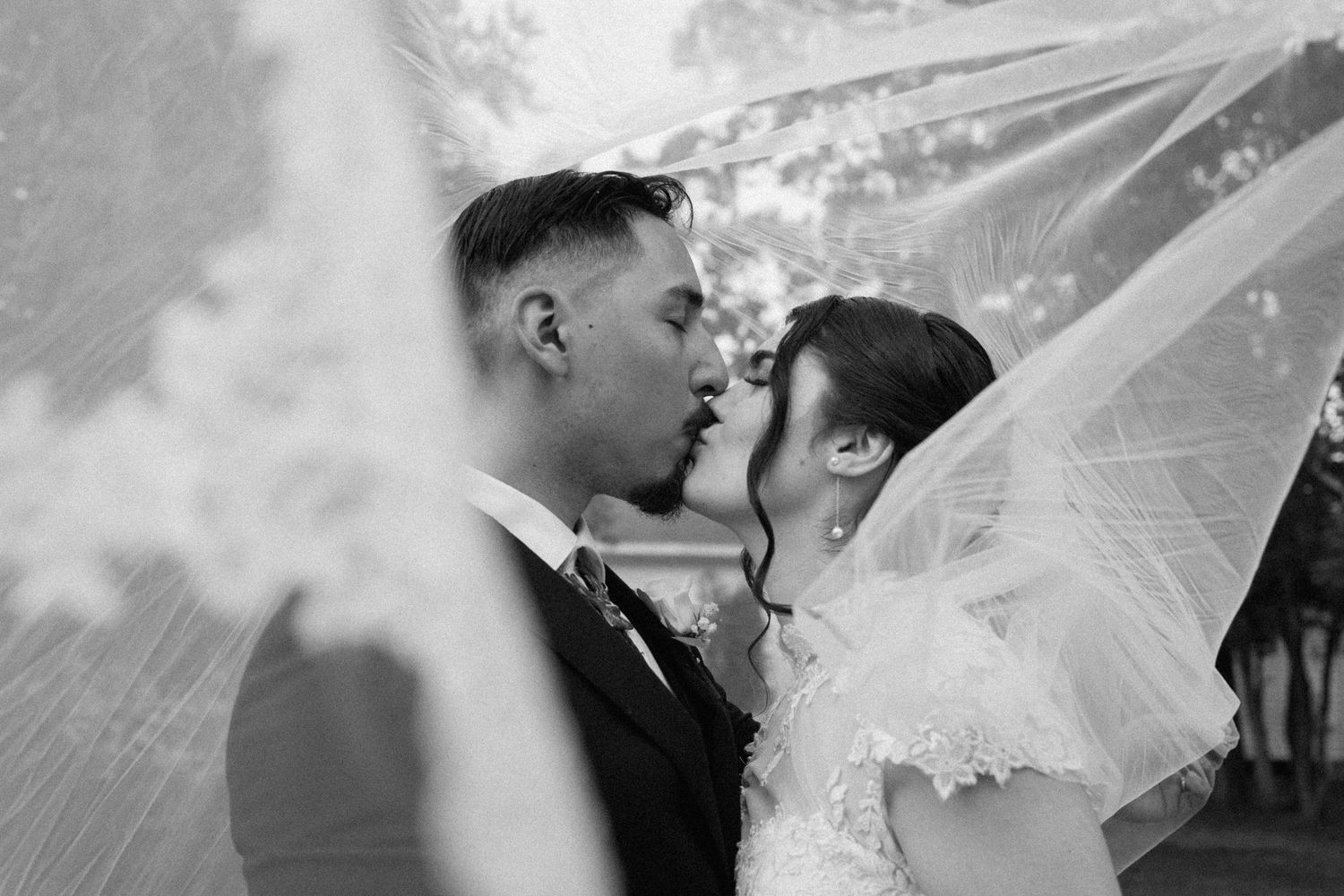 Black and white photo of bride and groom kissing under flowing wedding veil outdoors.