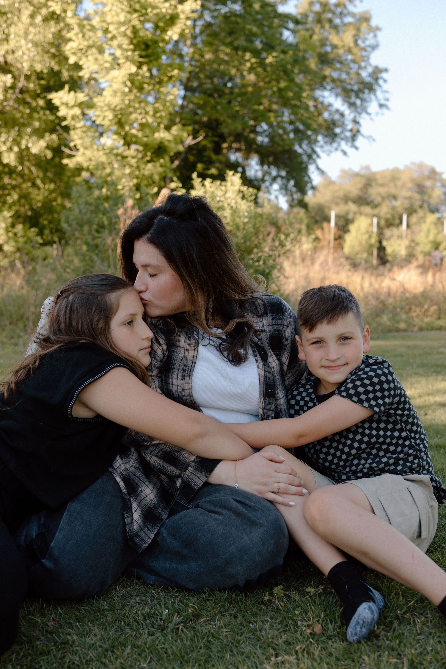 A mother sits on grass kissing her daughter while her son leans against her in a park.