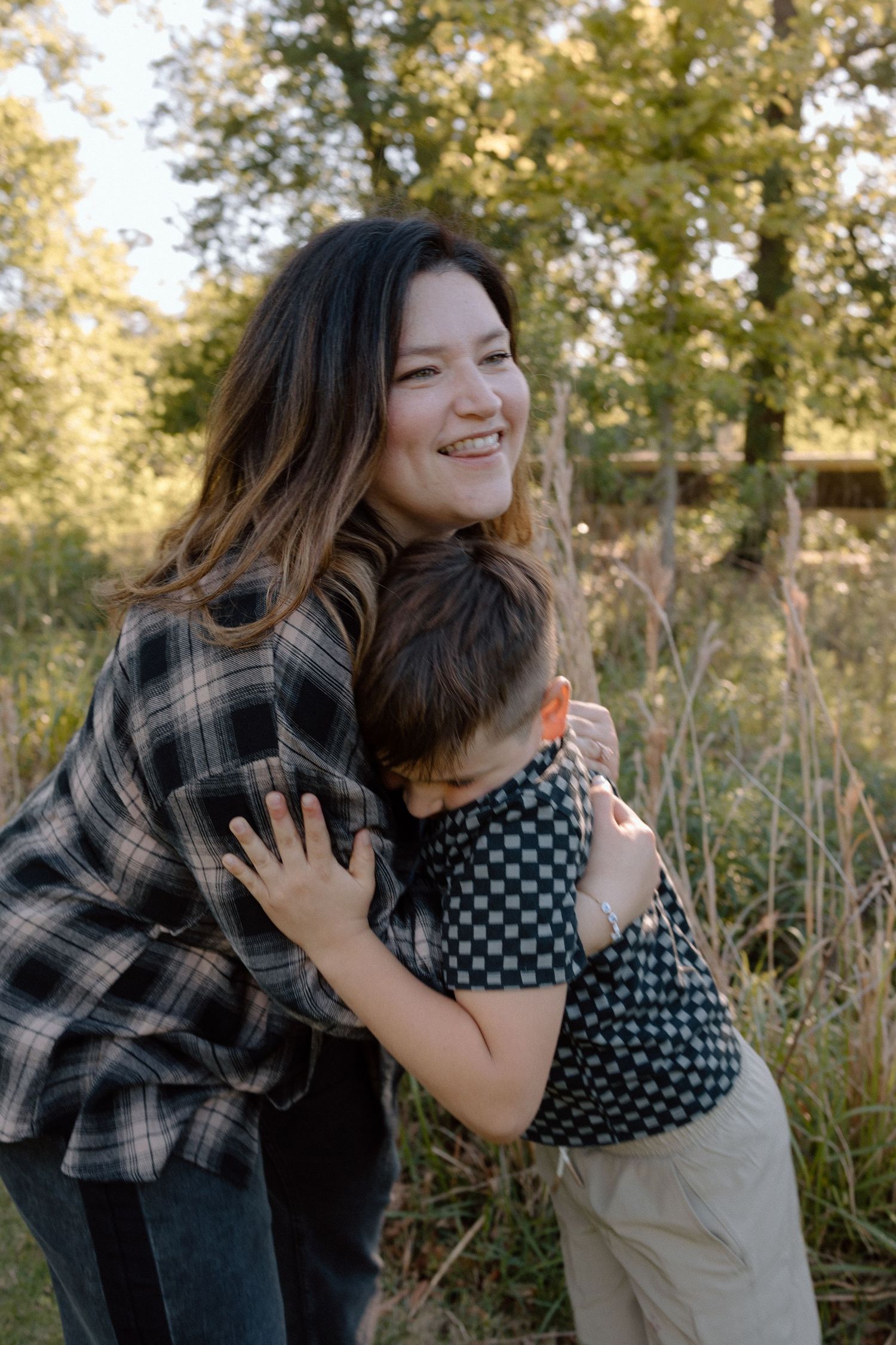 A smiling woman hugs a young boy outdoors in a sunlit forest setting.