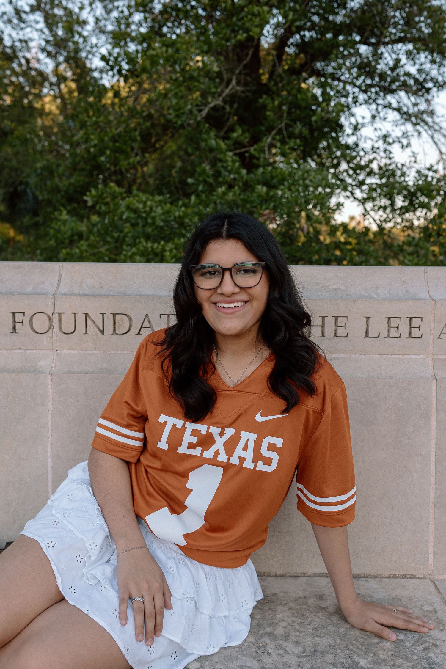 Smiling young woman in Texas Longhorns jersey and glasses sitting by stone foundation wall.