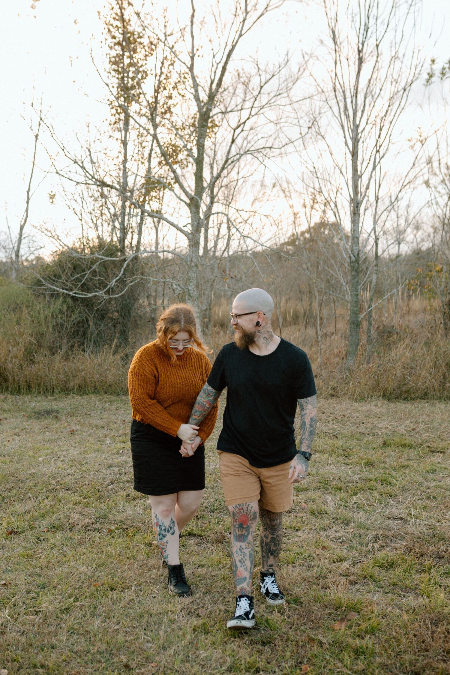 A tattooed couple holds hands and walks together through a field of bare trees in autumn.