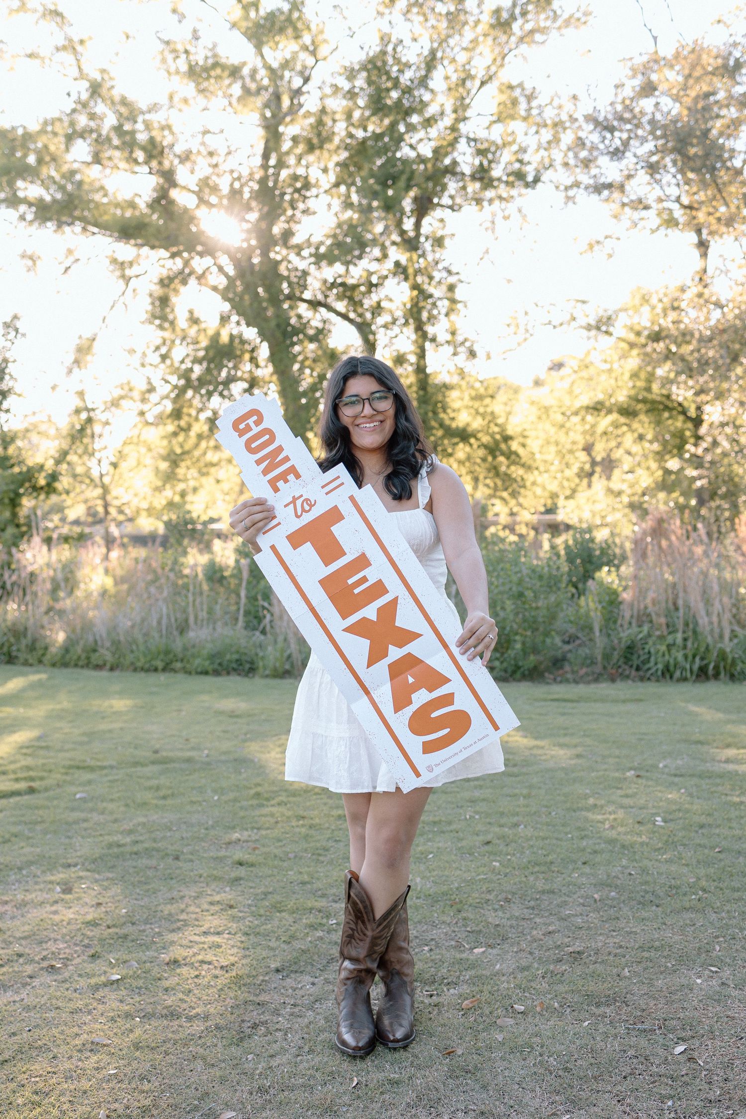 Smiling young woman in white dress and cowboy boots holding Gone to Texas sign outdoors.