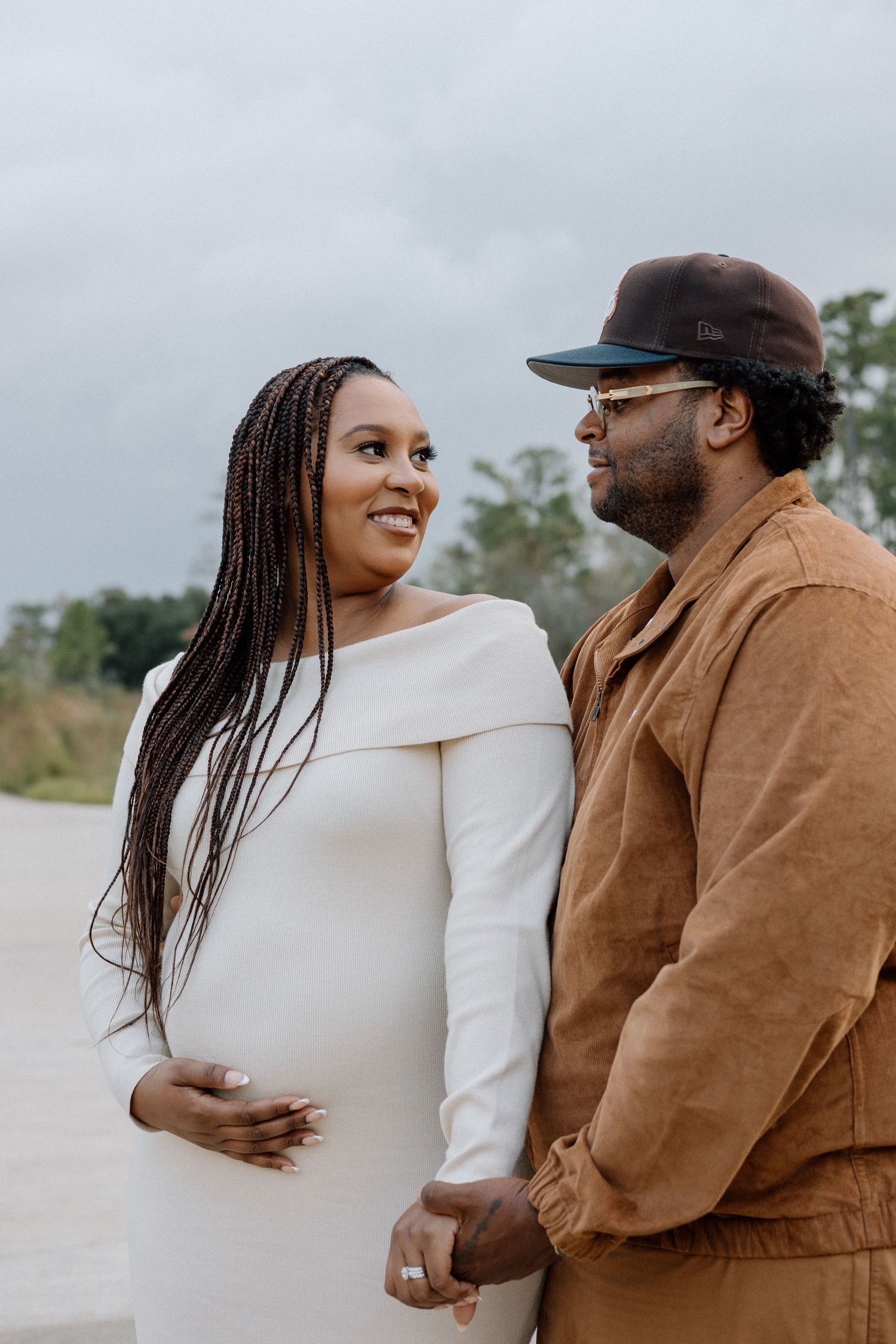Smiling pregnant woman in white dress holds hands with partner outdoors on cloudy day.