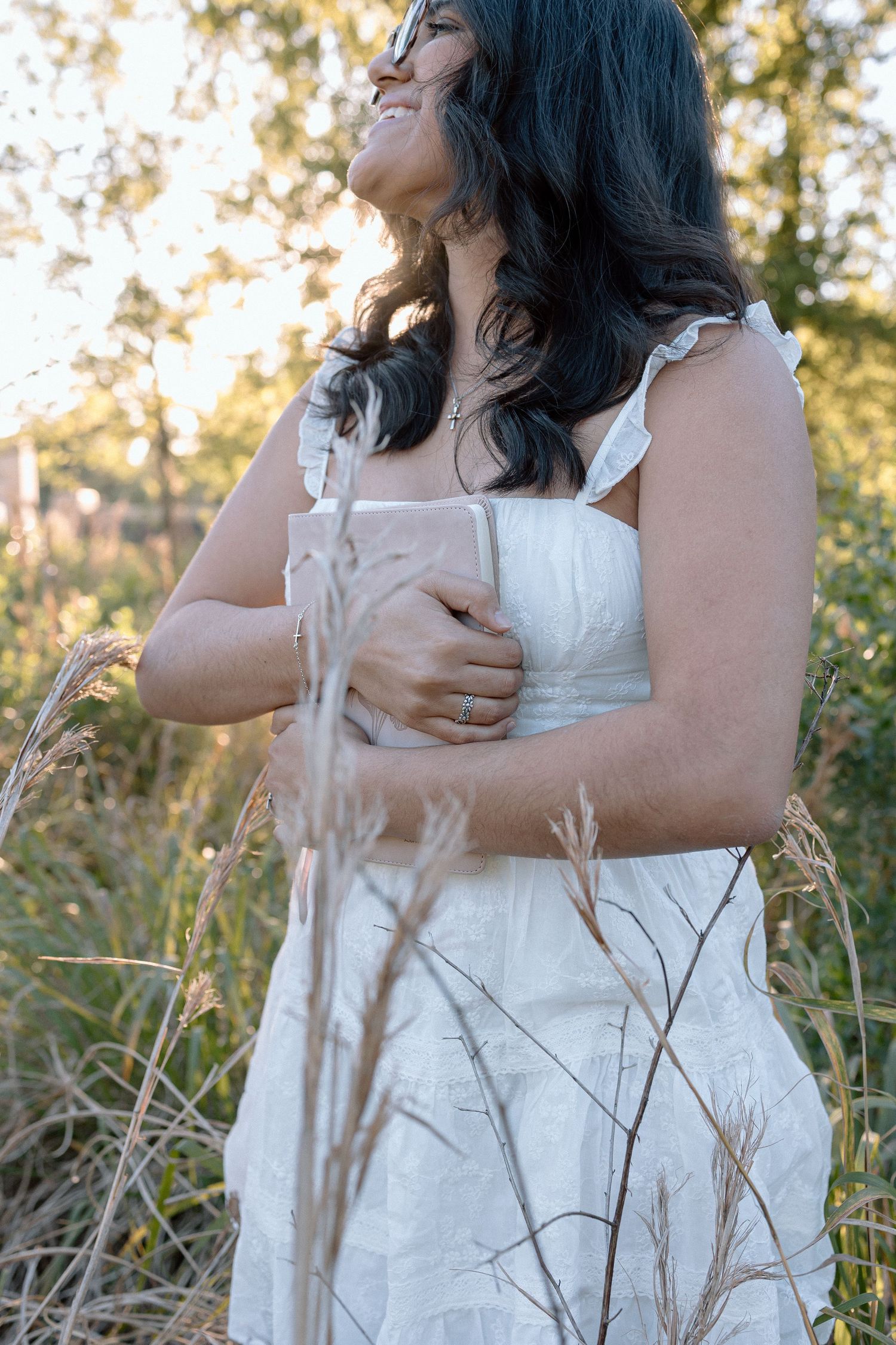 Smiling woman in white dress holding book among tall golden grasses in sunlit field.