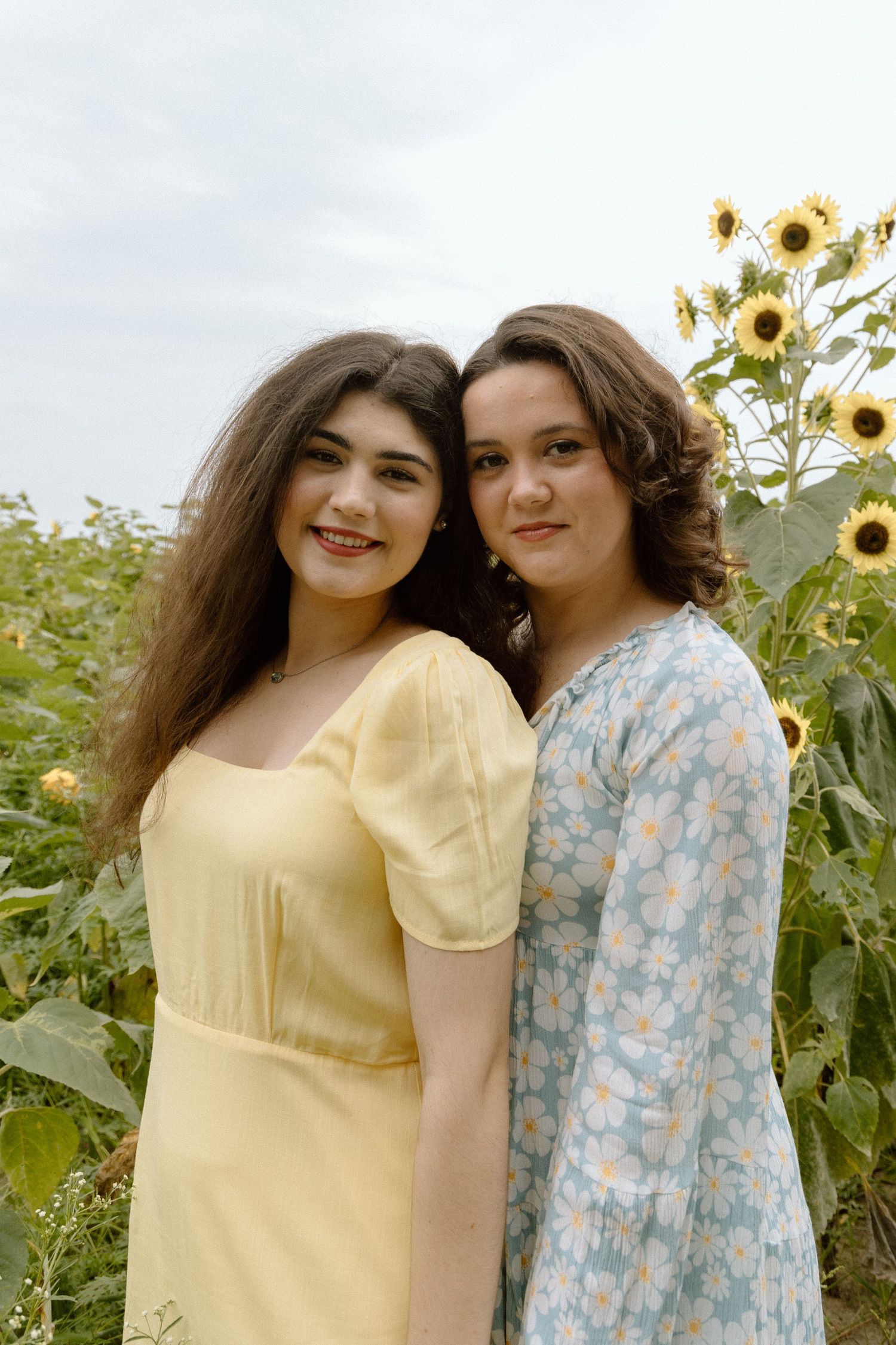 Two smiling young women in yellow and floral dresses posing together in a sunflower field.