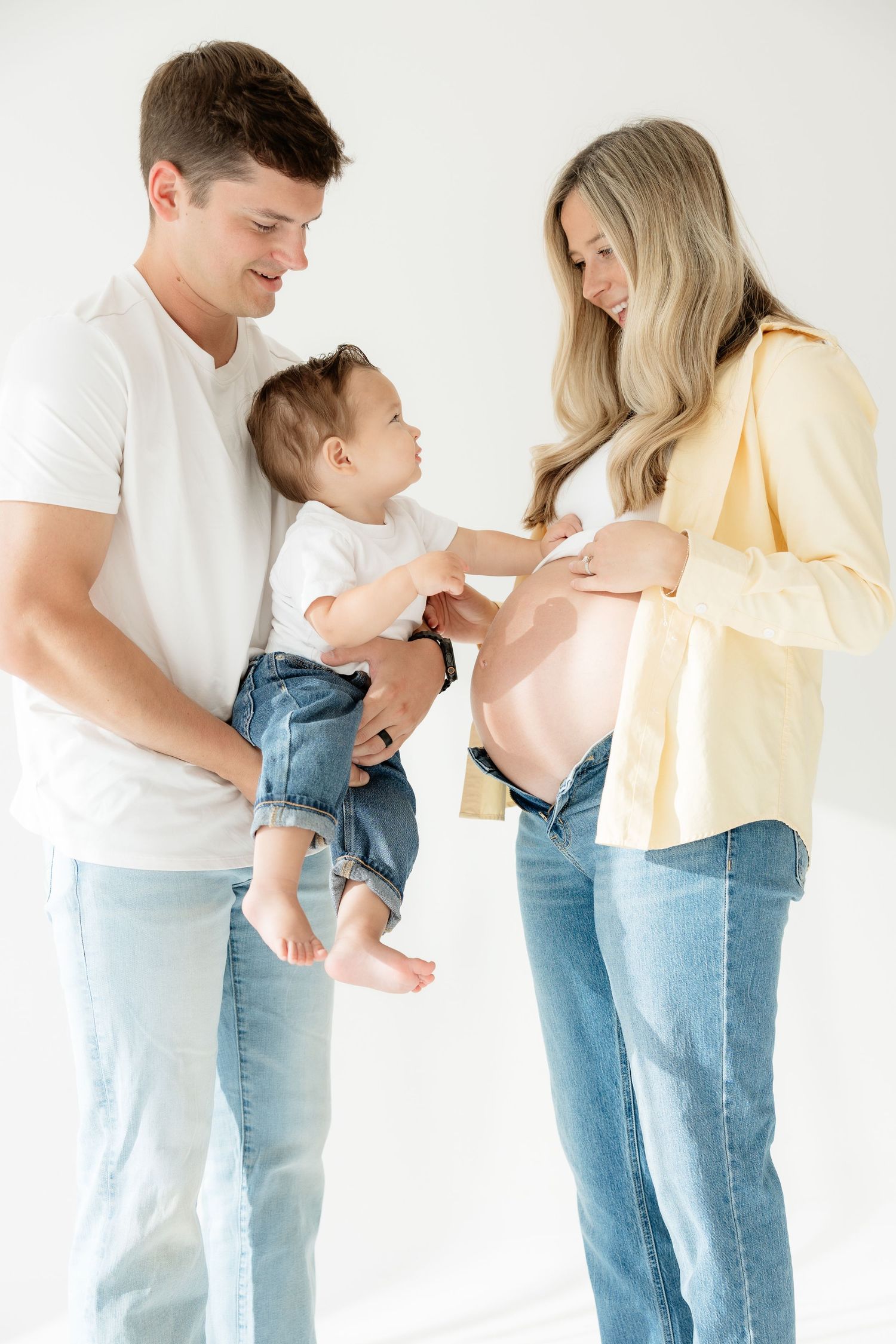 A smiling couple in jeans with a baby touching the pregnant mothers bare belly against a white background.