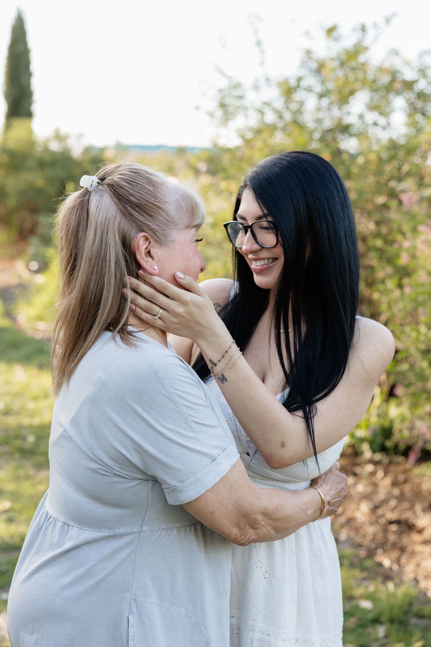 Two women embrace outdoors, one smiling in white dress with glasses, the other in light blue, surrounded by greenery.