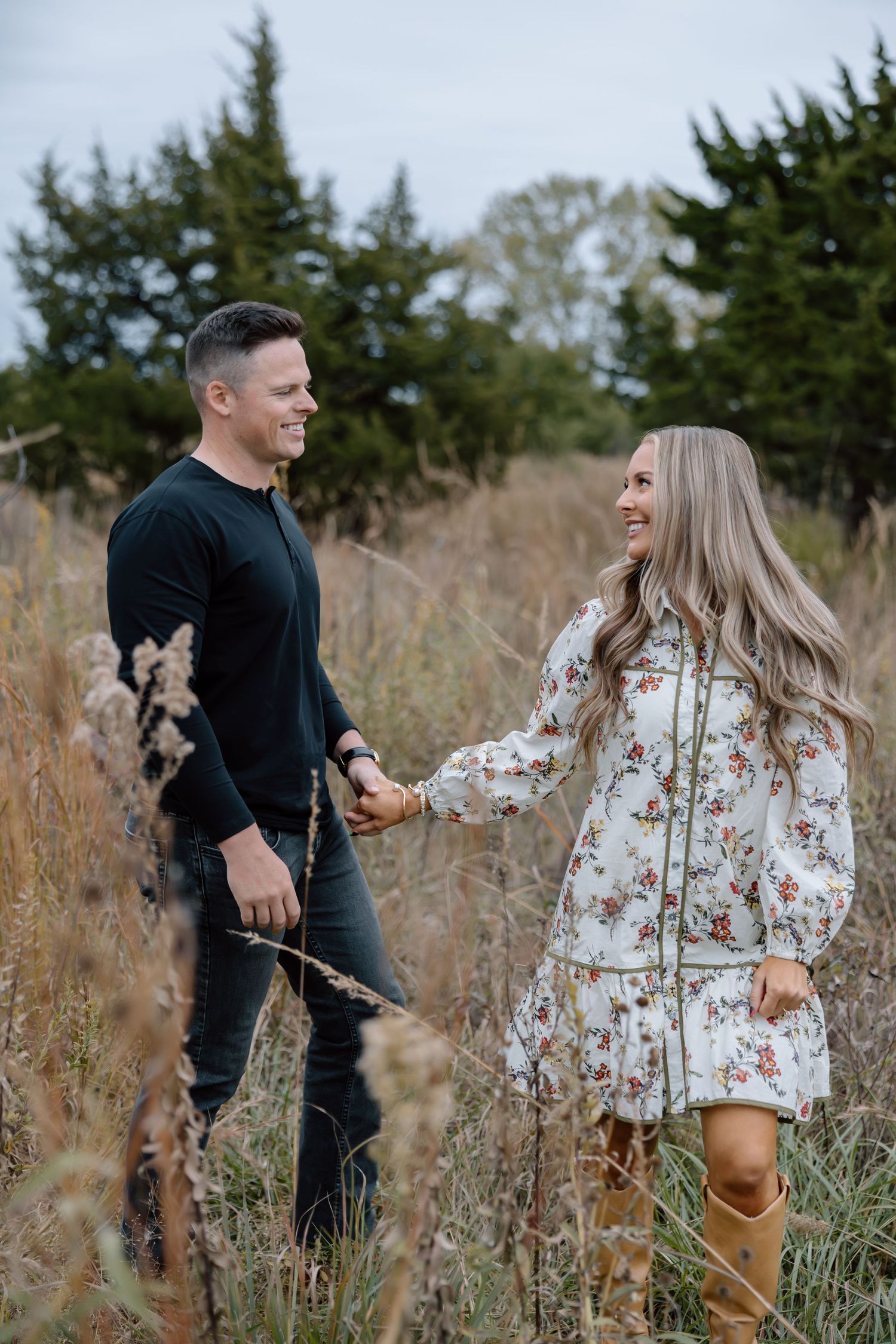 Happy couple holding hands and smiling in a golden autumn meadow with evergreens.